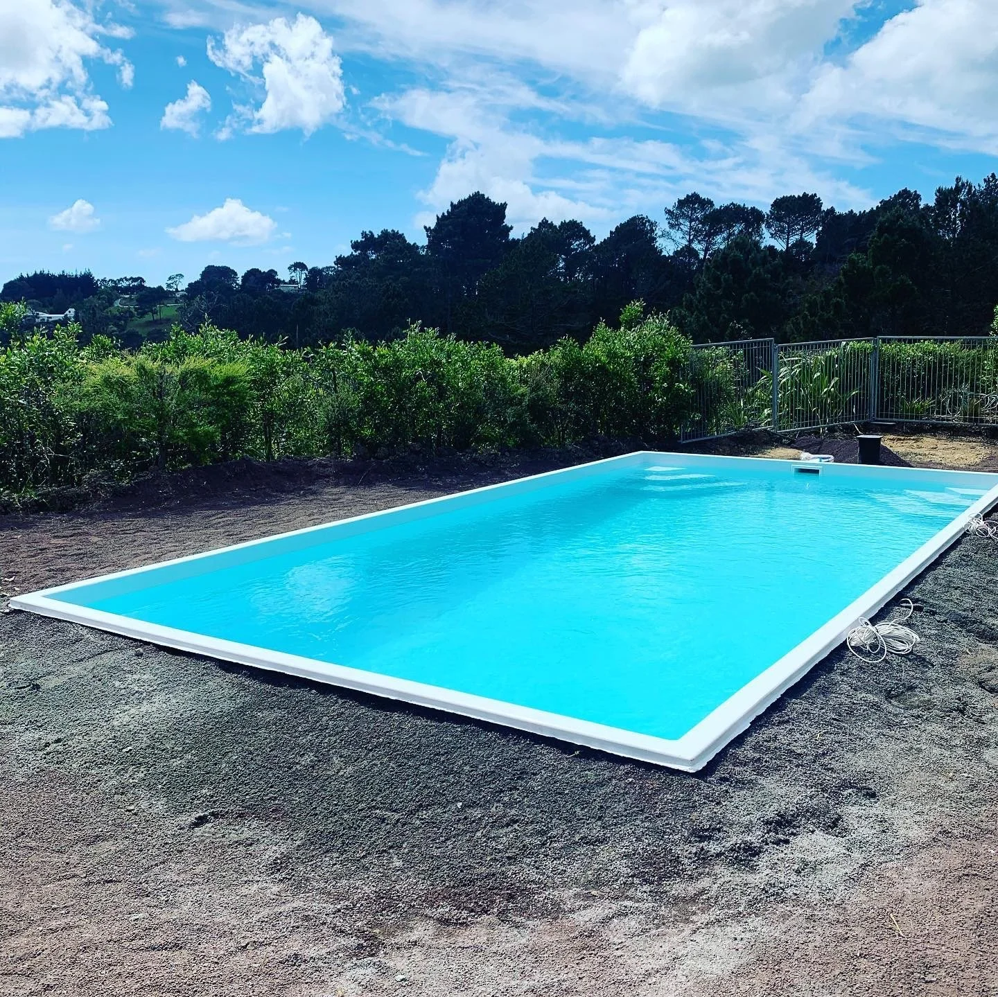 Unempty rectangular above-ground swimming pool filled with water, situated outdoors on dirt ground, with a background of green bushes, trees, and a partly cloudy sky.