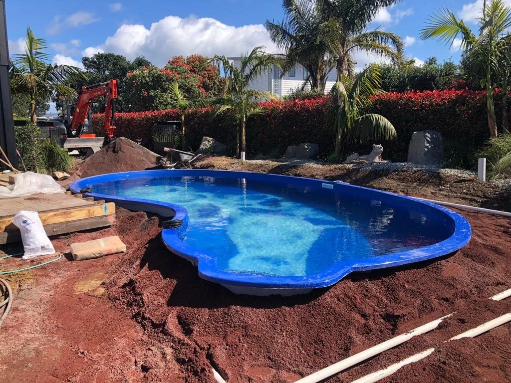 In-ground swimming pool under construction with surrounding dirt, construction equipment, and tropical plants.