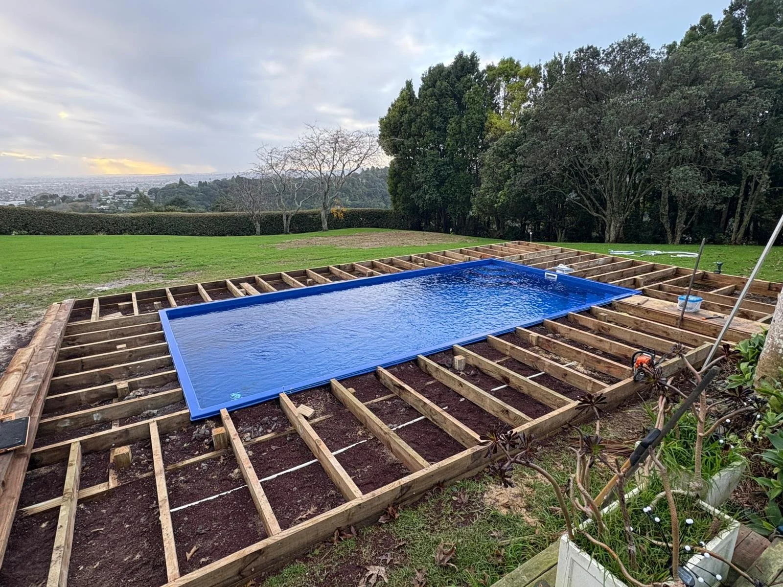 A swimming pool with a bright blue liner is under construction in a backyard with wooden framing, surrounded by a grassy lawn and trees in the background.