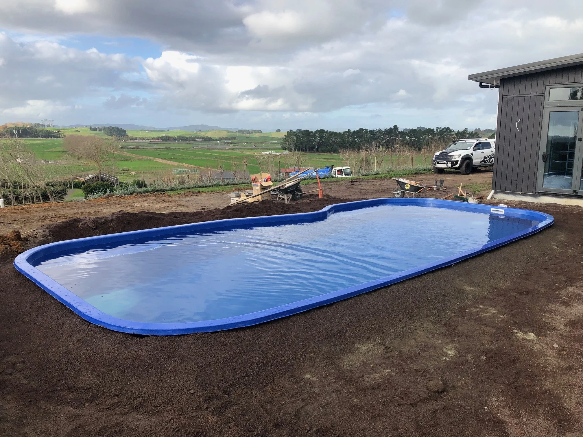 In-ground swimming pool with a blue border on dirt ground, outside of a house, with a scenic rural landscape in the background and a few parked vehicles nearby.