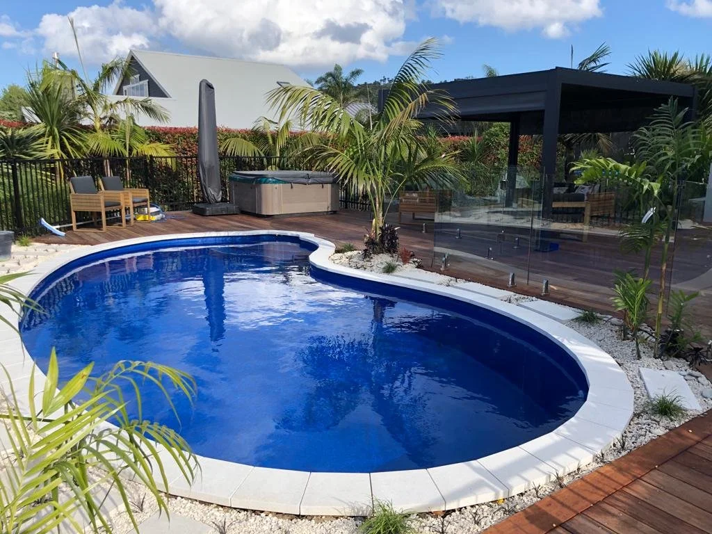 Empty backyard swimming pool surrounded by white tiles, lush tropical plants, and outdoor furniture, with a wooden deck and a hot tub in the background.