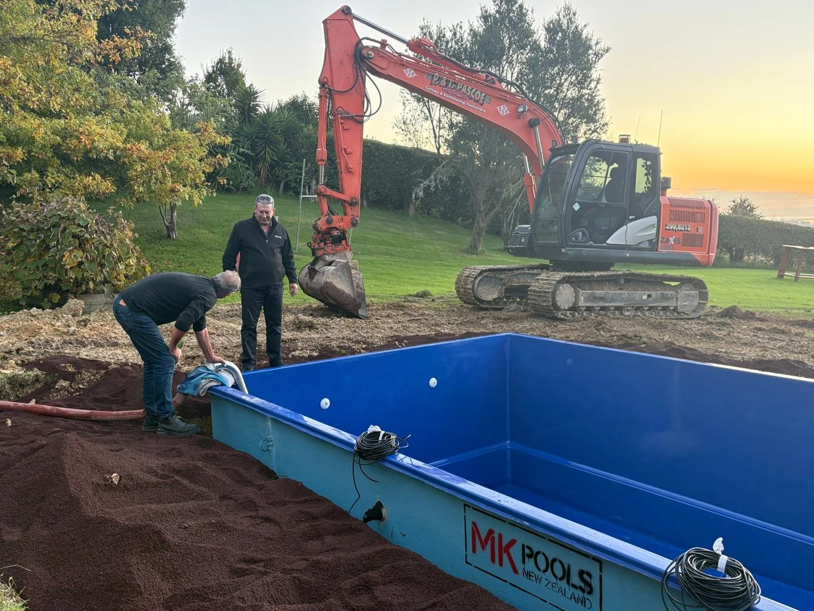 Two men are installing a swimming pool, with one man bending down to connect accessories while the other watches. There is an excavator behind them on a grassy area during sunset, with trees and bushes in the background.