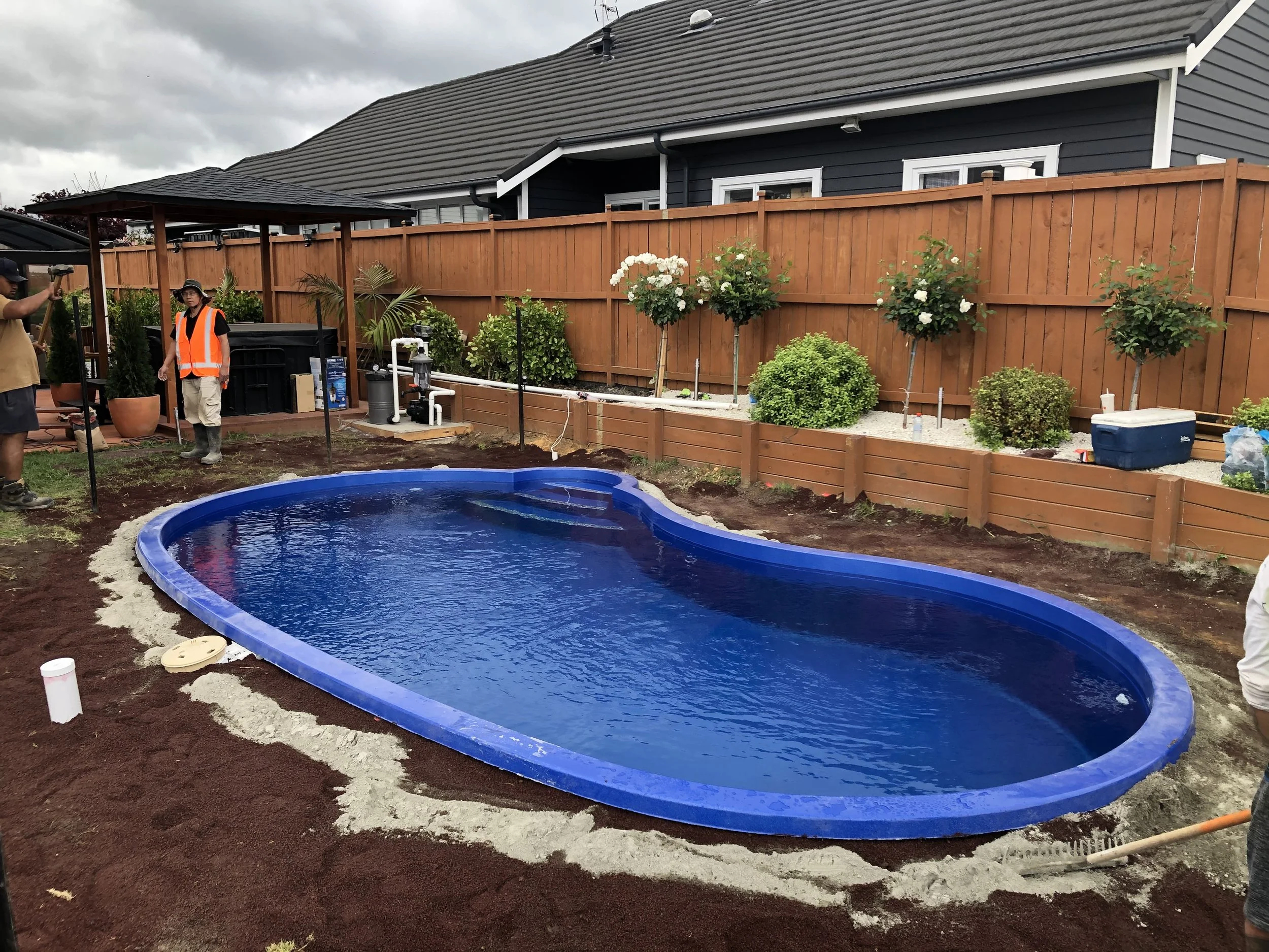 An in-ground swimming pool under construction in a backyard with trees and a wooden fence, with workers present.