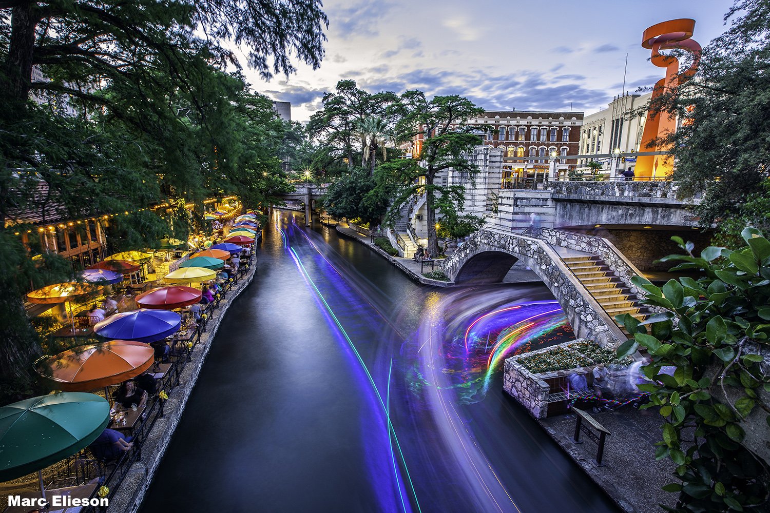 A vibrant scene of the San Antonio River Walk during evening, with colorful umbrellas shading outdoor dining tables along the riverbank, and blurred moving boats creating light trails on the water, framed by lush trees and historic buildings.