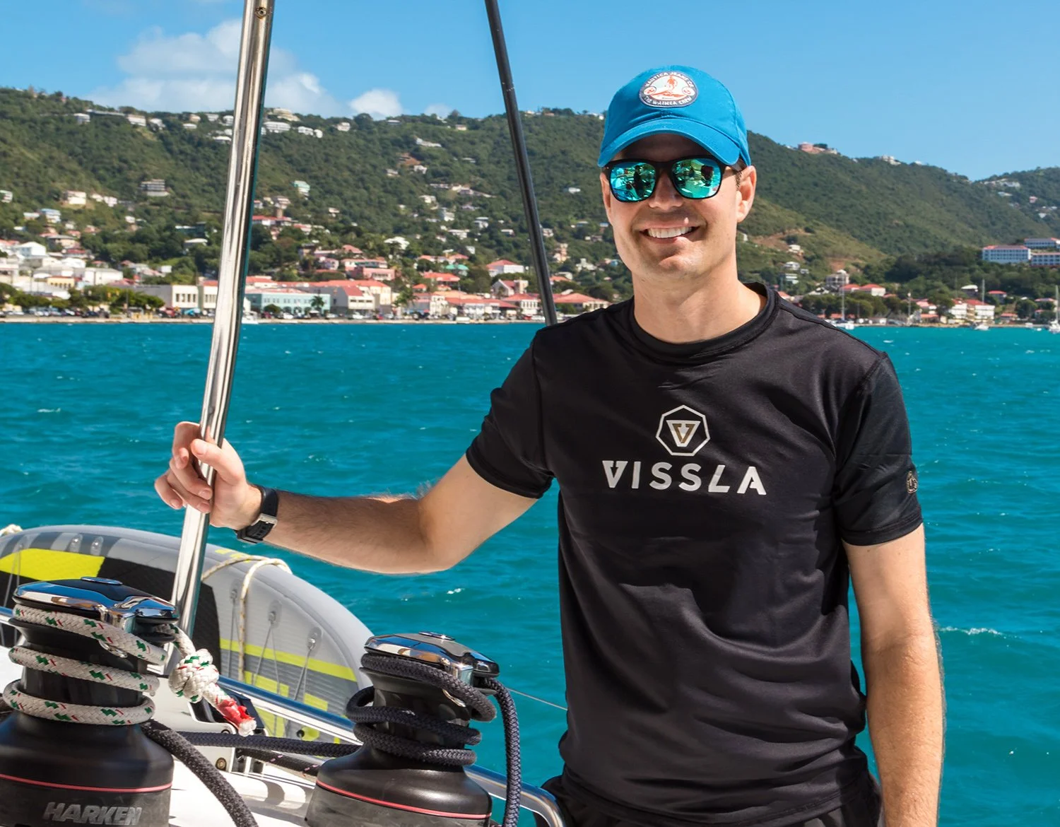 Man on a boat smiling, wearing sunglasses, a blue cap, and a black T-shirt with 'VISSLA' logo, with a coastal town and hillside in the background.