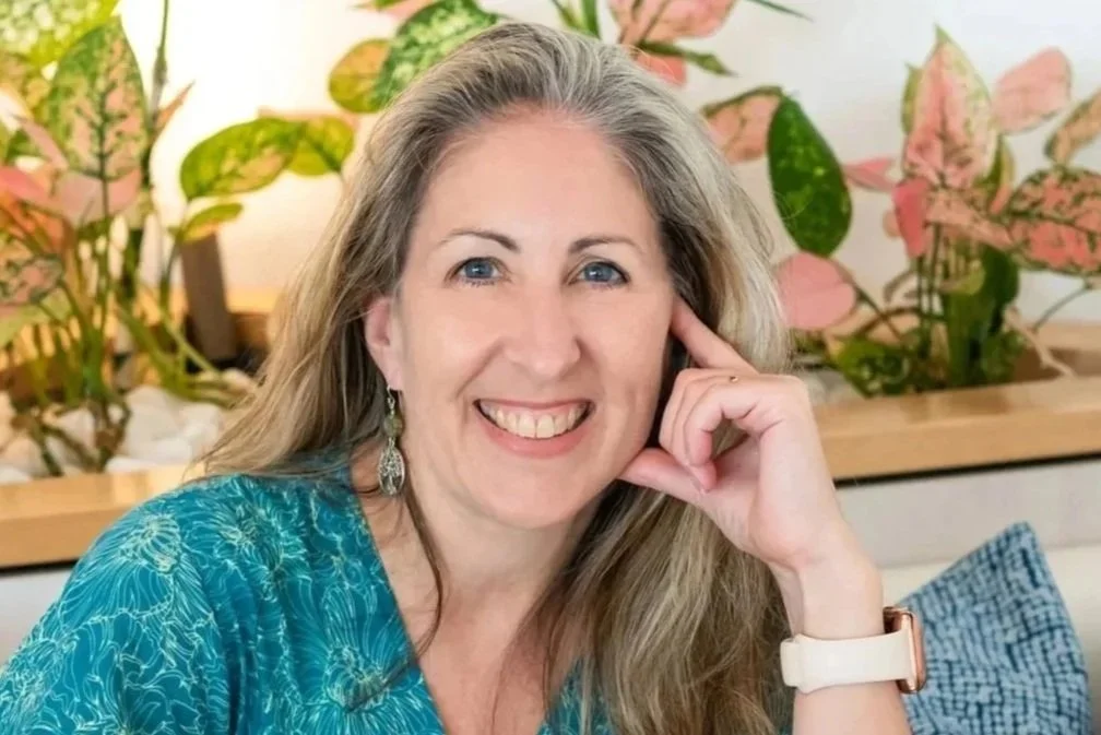 A smiling woman with long blonde hair and blue eyes, wearing a turquoise patterned top, silver earrings, and a white watch, sitting indoors with pink and green plants in the background.