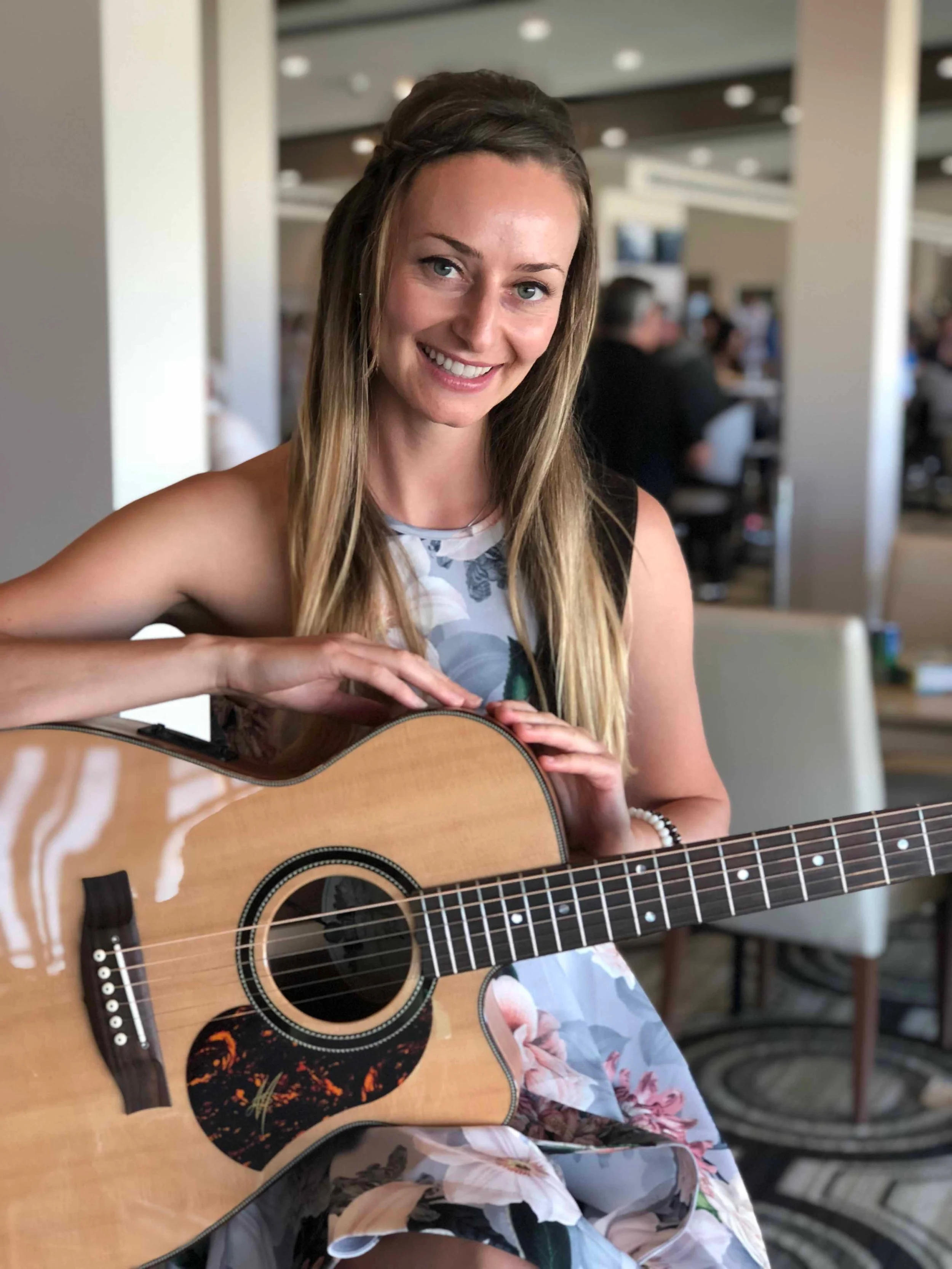 A woman smiling and holding an acoustic guitar indoors at a social event or restaurant.