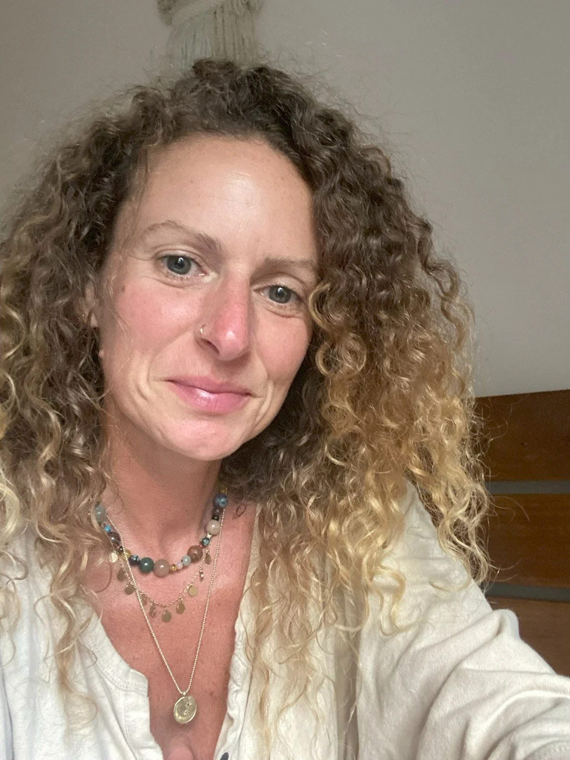 A woman with curly blonde hair, wearing jewelry, taking a selfie indoors.