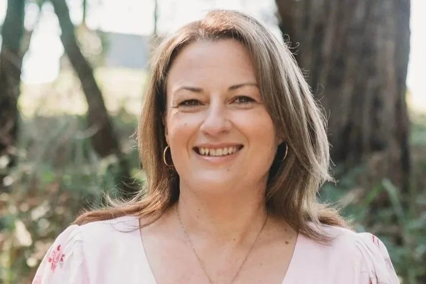 A woman with light brown hair smiling outdoors, wearing a white top and hoop earrings, with trees and greenery in the background.