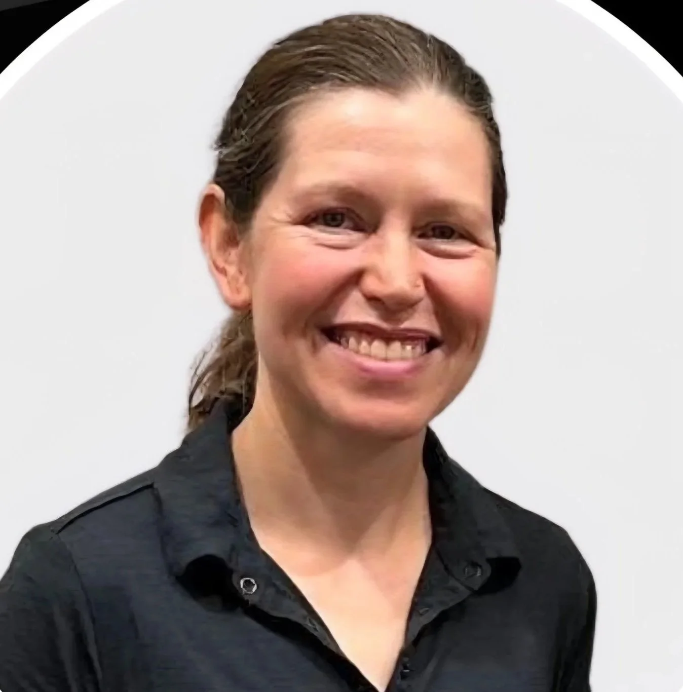 A woman with brown hair tied back, smiling, wearing a black collared shirt, against a plain white background.