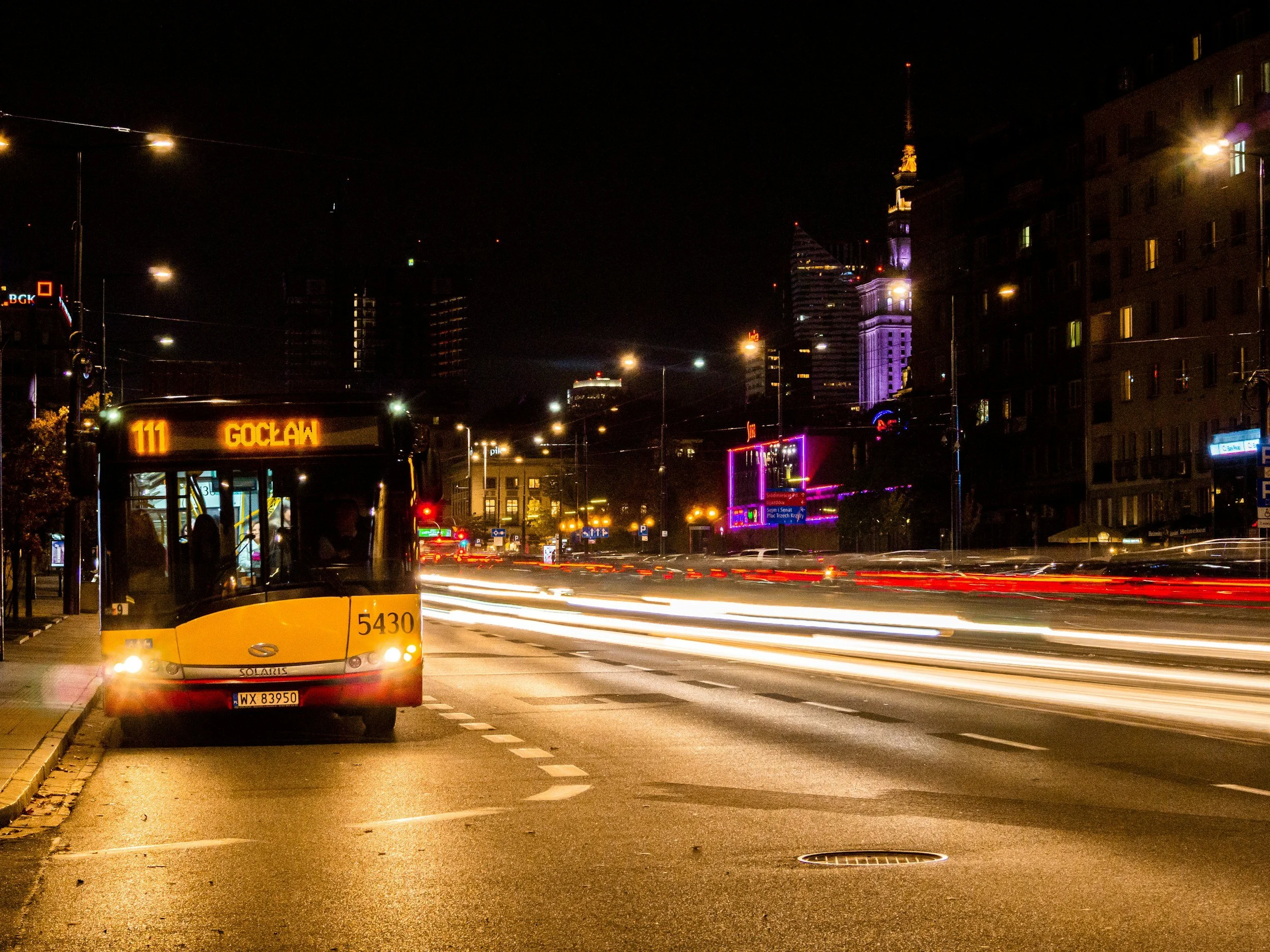 Night city street with moving cars and a bus labeled 'GOCŁAW', illuminated buildings, and light trails from traffic.