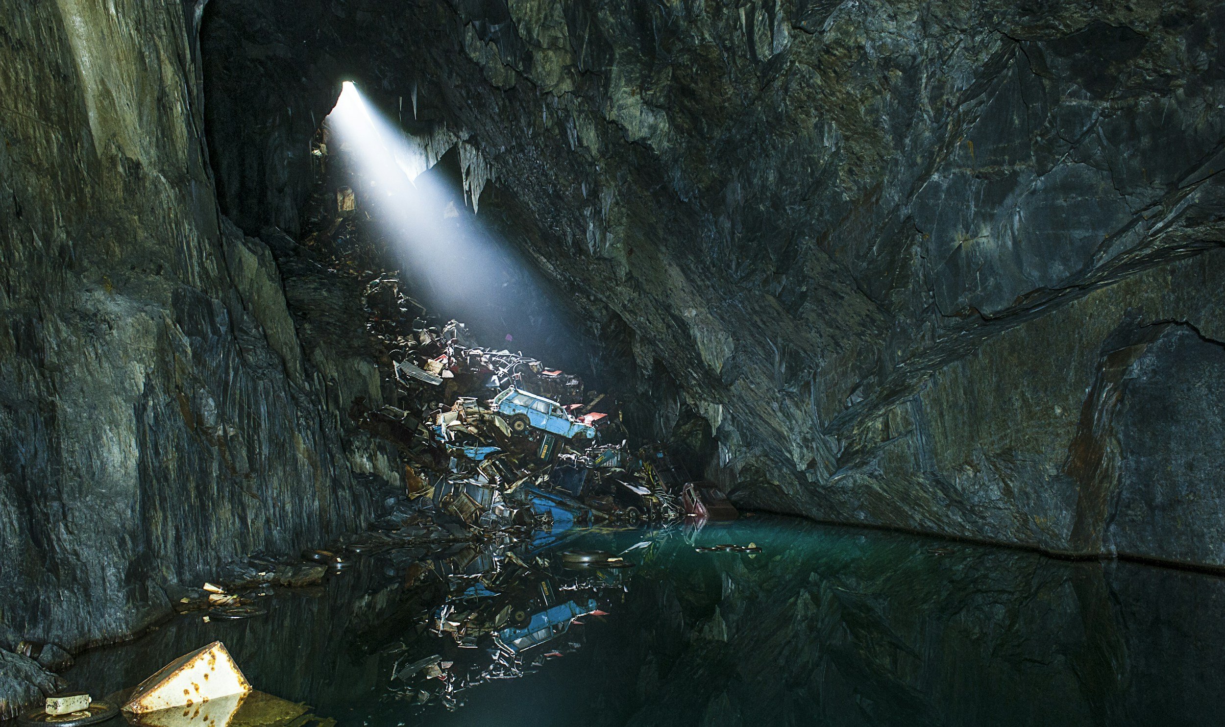 Inside an underground cave with a small pool of water, a pile of discarded old cars and debris, and a beam of light shining through an opening at the top.