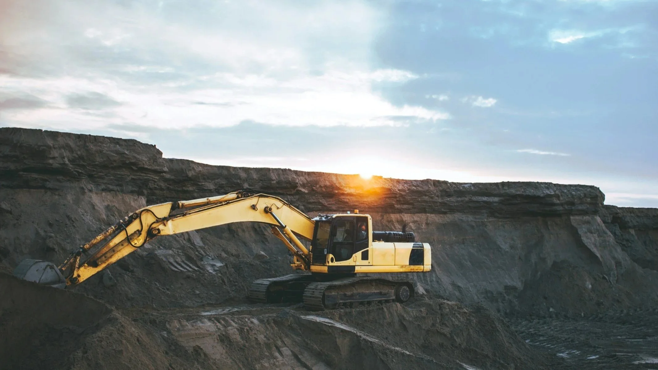 Construction site with two yellow excavators working on dirt, near a cliff at sunset.