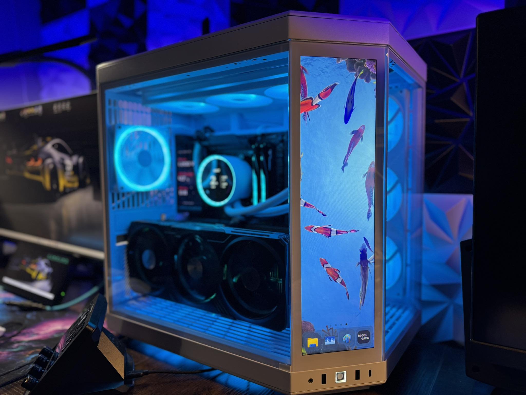 A gaming computer with a transparent side panel showing internal components, illuminated by blue LED lights, with a smartphone screen displaying a fish wallpaper in the foreground.