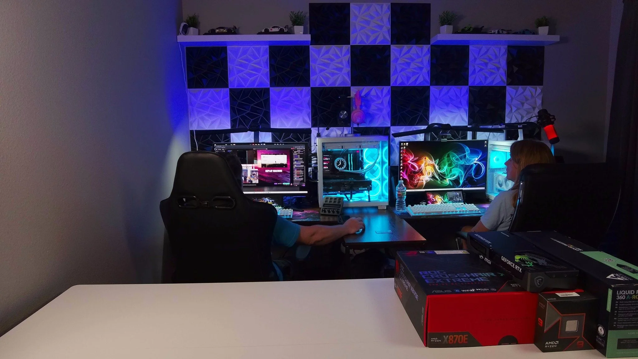 A gaming setup with three monitors, colorful LED lighting, and two people sitting at the desk. The wall behind has decorative acoustic panels and shelves with small models or toys. Several boxes of computer hardware are on a white table in the foreground.