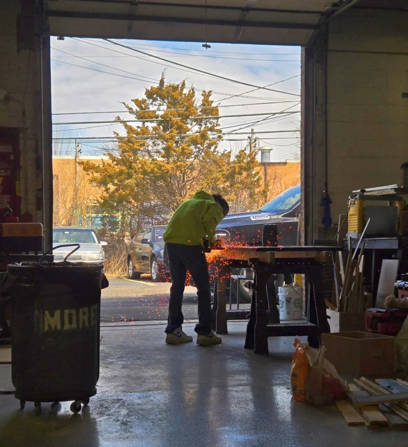 A person wearing a yellow hoodie and dark pants is welding in a garage, sparks flying from the work. Outside, there are parked cars and a tree with autumn-colored leaves, visible through the garage open door.