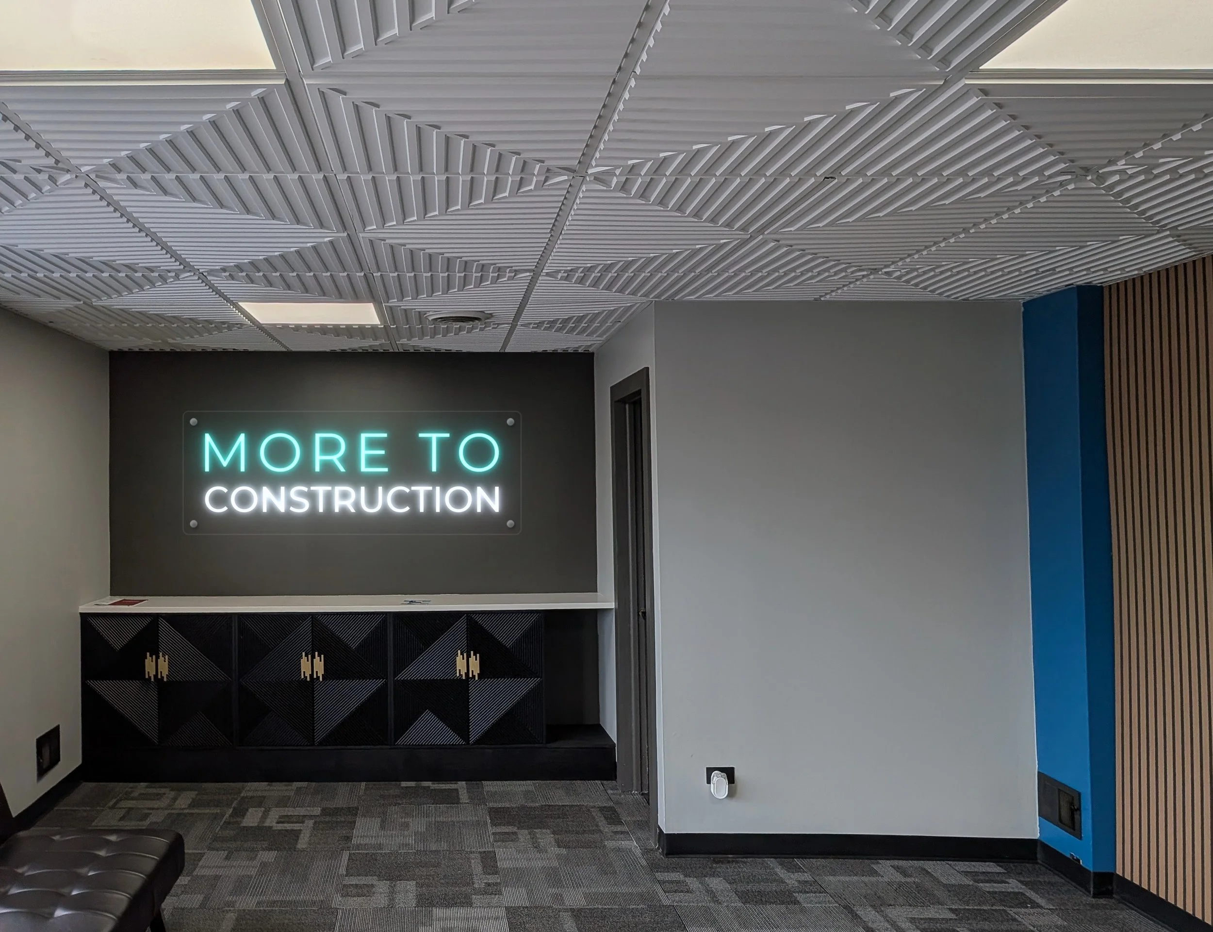 Interior space with a textured white ceiling, dark front desk with gold accents, and a sign that says 'MORE TO' in neon lights, with parts of a blue wall and a black leather couch visible.