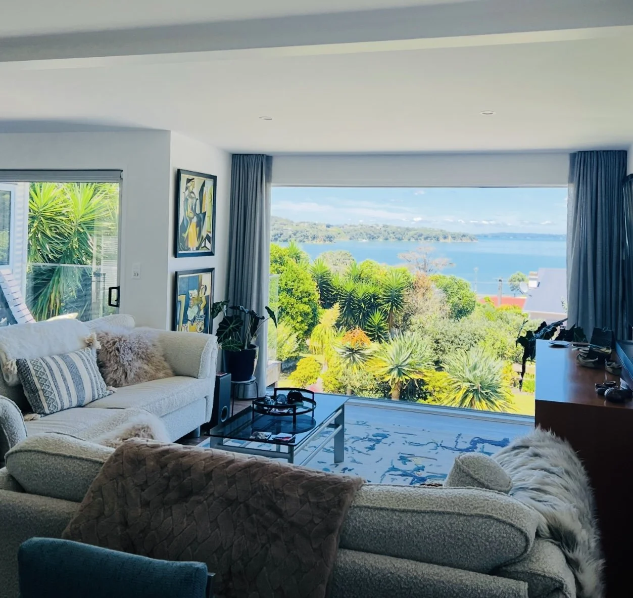 Living room with large window overlooking lush greenery and water, modern art on wall, cream-colored sofas, plants, and a coffee table.