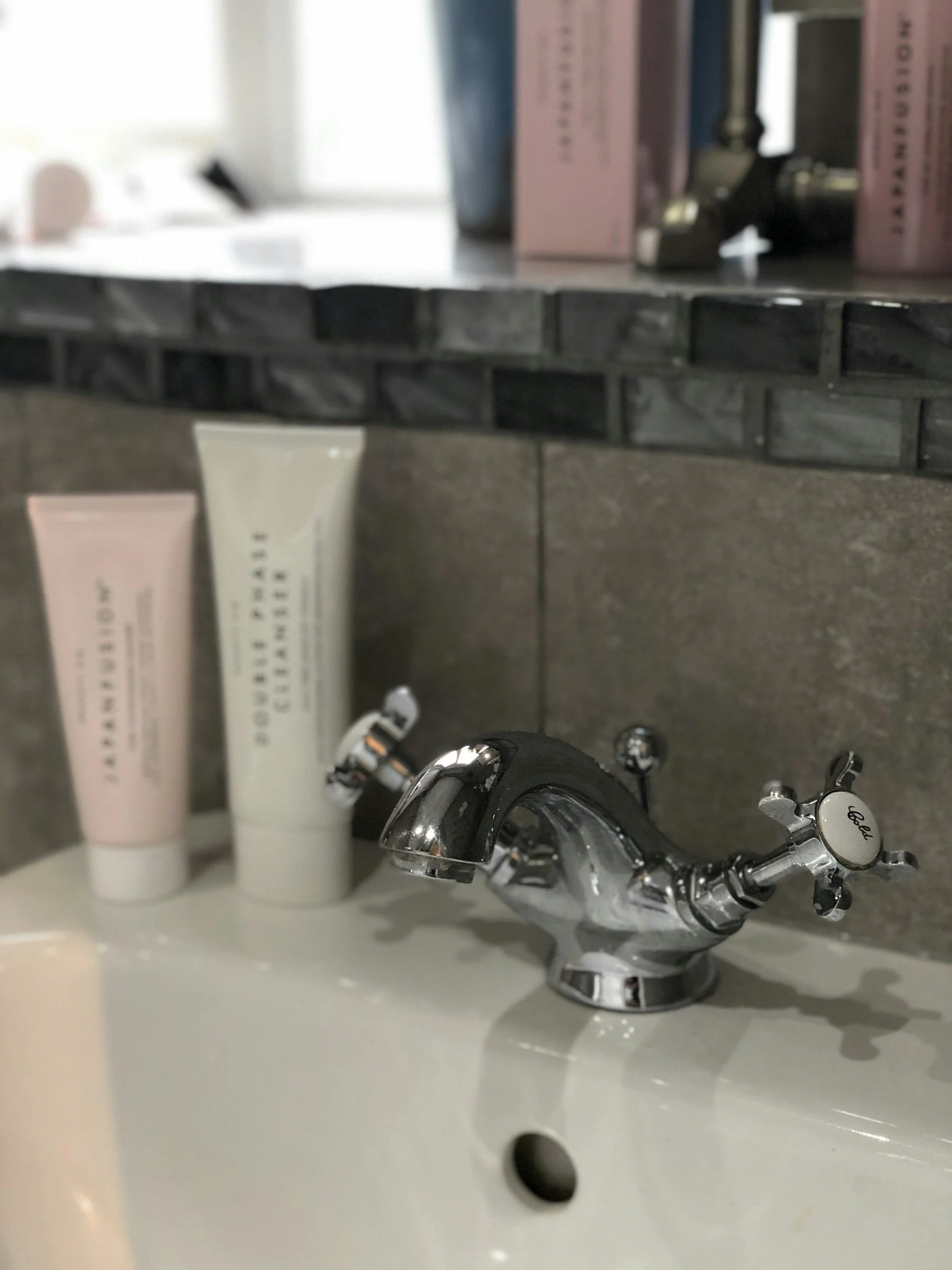 Bathroom sink with a chrome faucet, pink and white skincare tubes, and a backdrop of dark neutral tile with a mirrored shelf with beauty products.