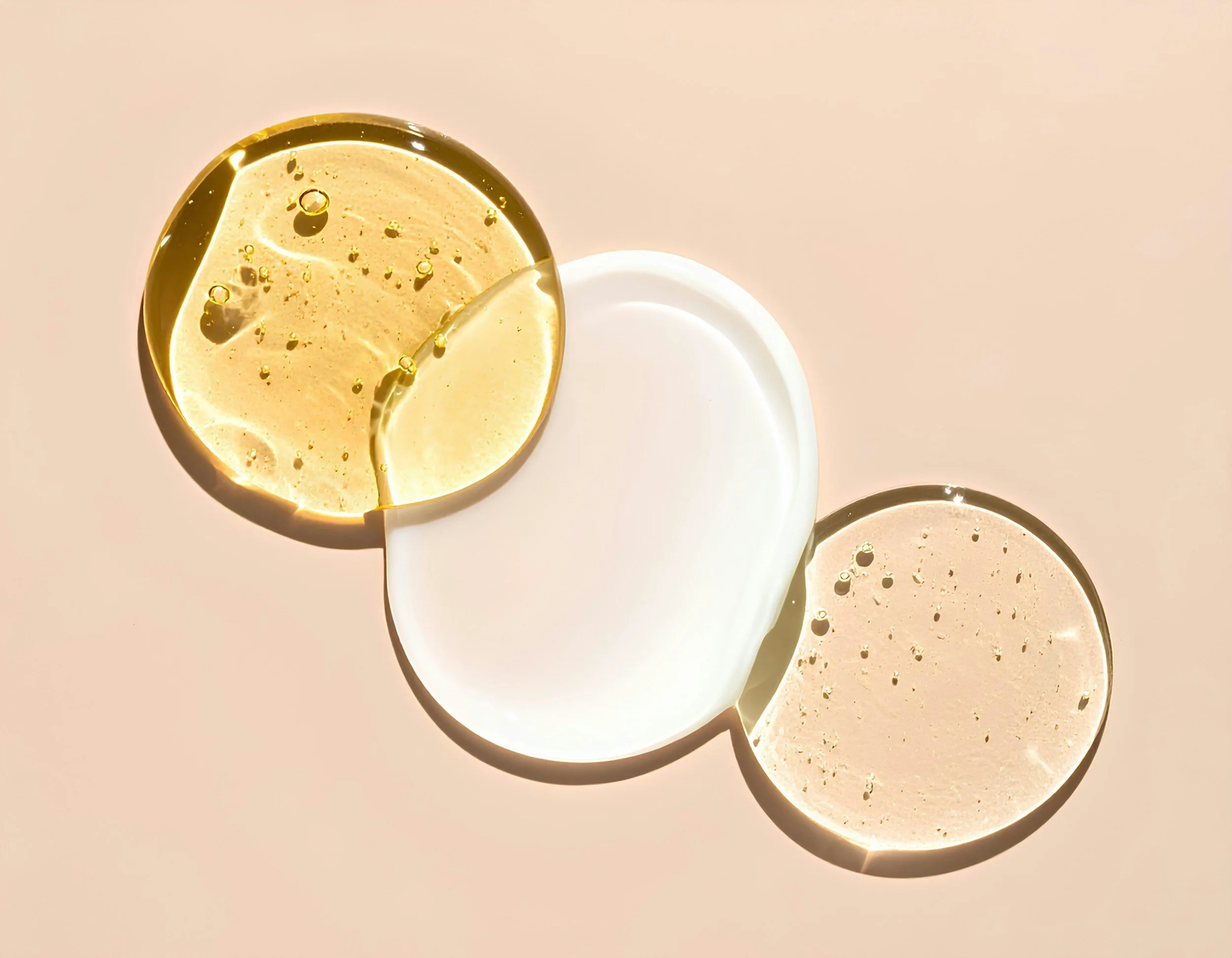 Three circular drops of fluid. One drop is golden, like oil. One drop is white, like lotion. The last drop is clear, like aloe vera. The lotion is in the middle and overlaps the other two droplets, placed on a light peach background.