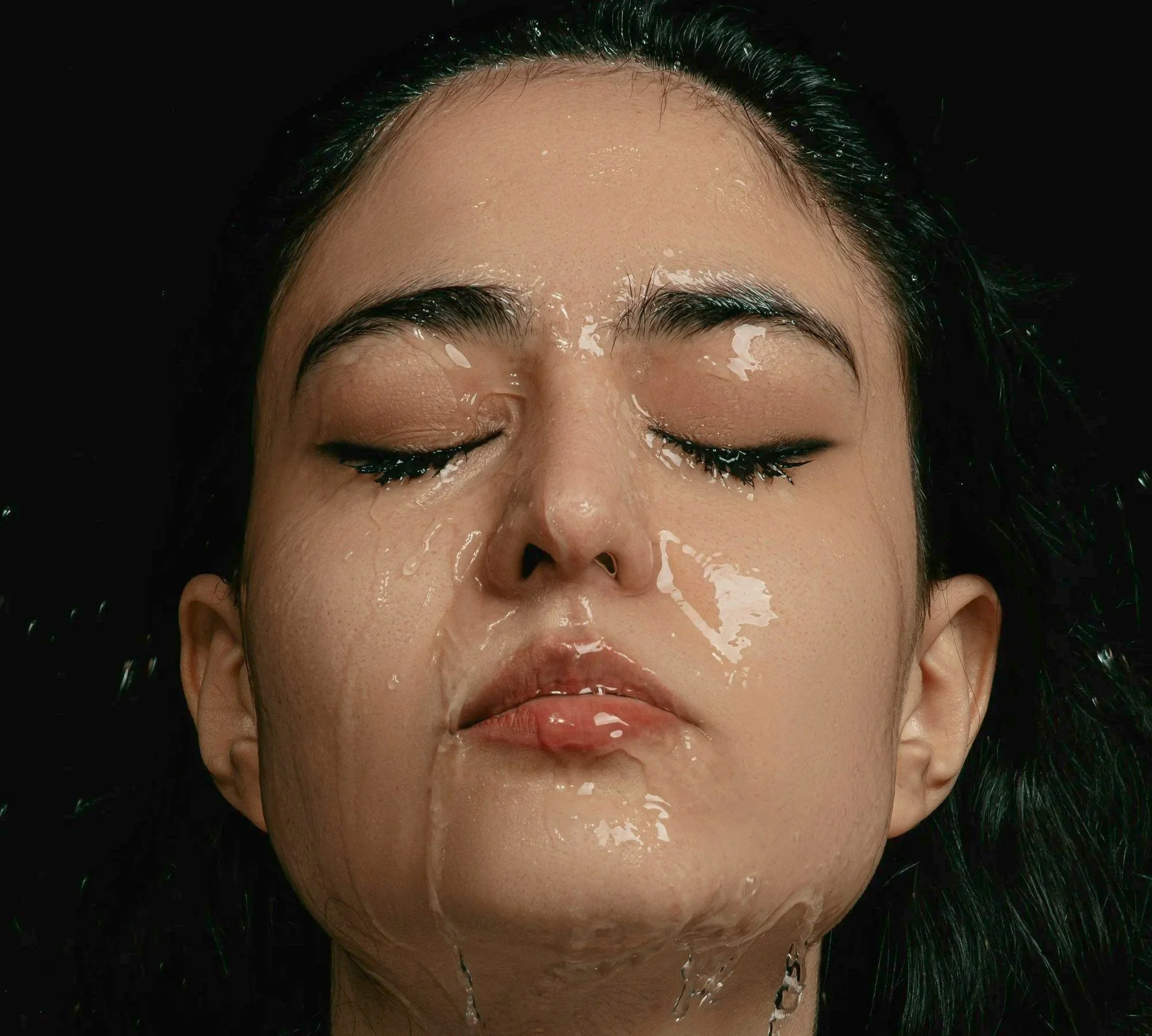A woman with black hair, thick eyebrows, and makeup has her eyes closed and water pouring down her face against a black background.