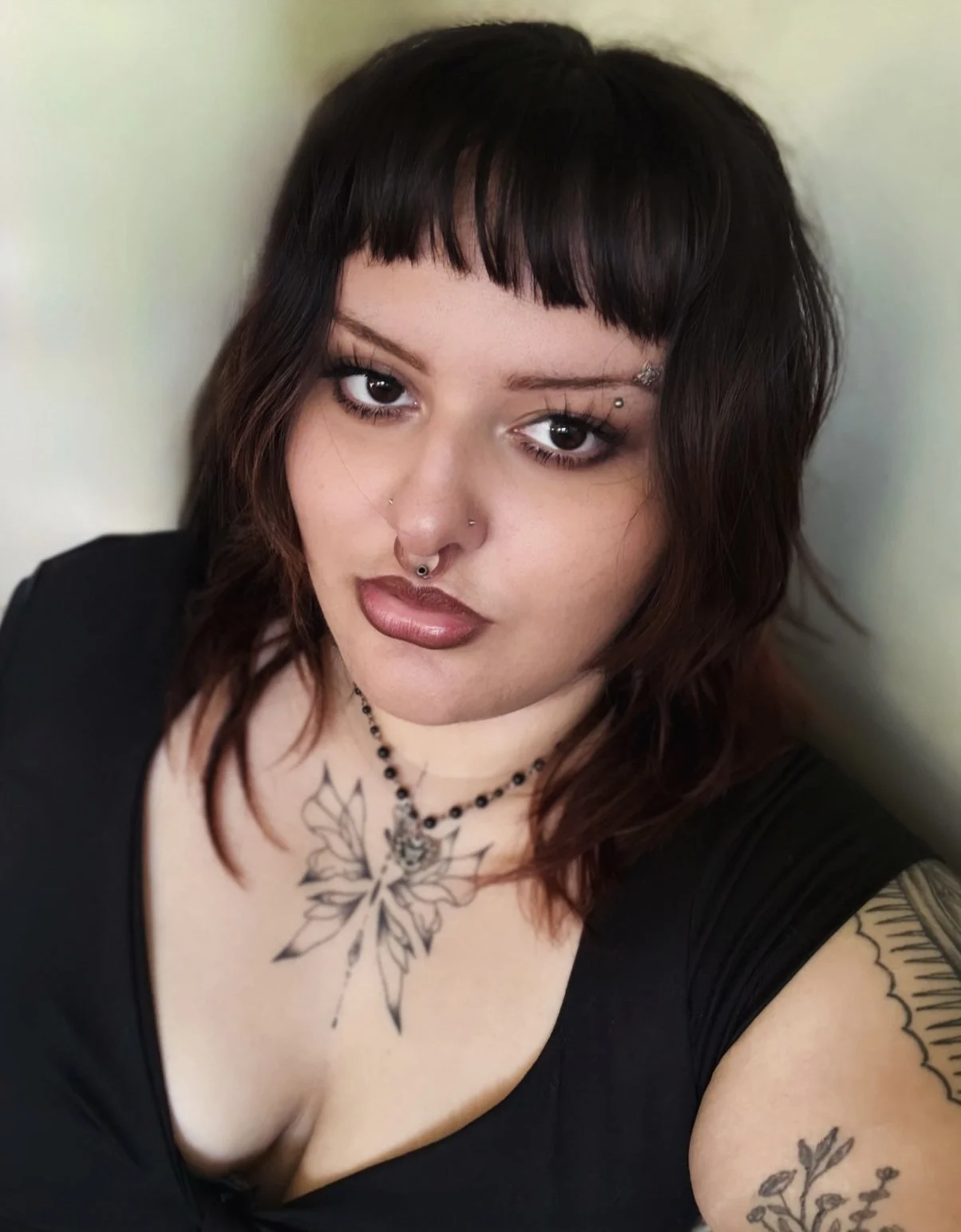 A Hispanic goth woman with dark hair, tattoos, and piercings taking a selfie against a light-colored wall.
