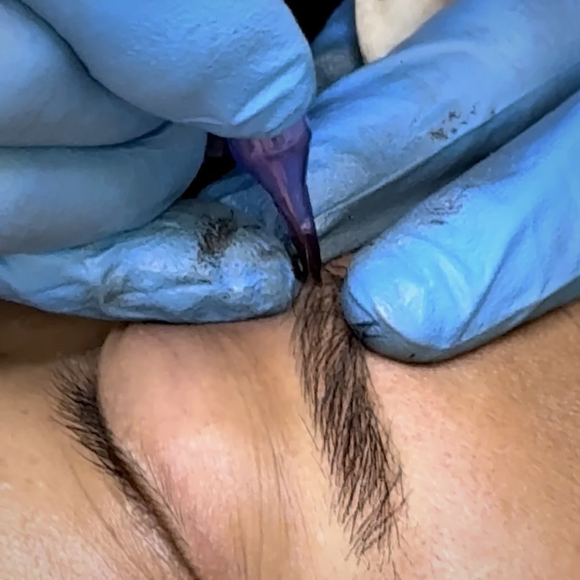 A close-up of nano brows machine hair strokes permanent makeup tattoo being created on a woman's eyebrow with a tattoo machine, held by a person wearing blue gloves.