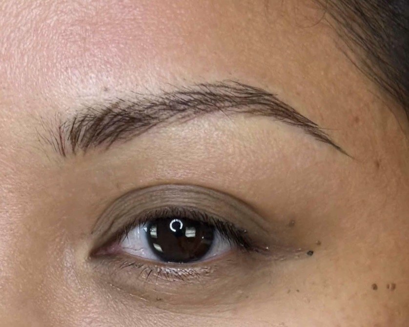Close-up of a person's eye and tattooed eyebrow, with visible skin texture and small freckles.