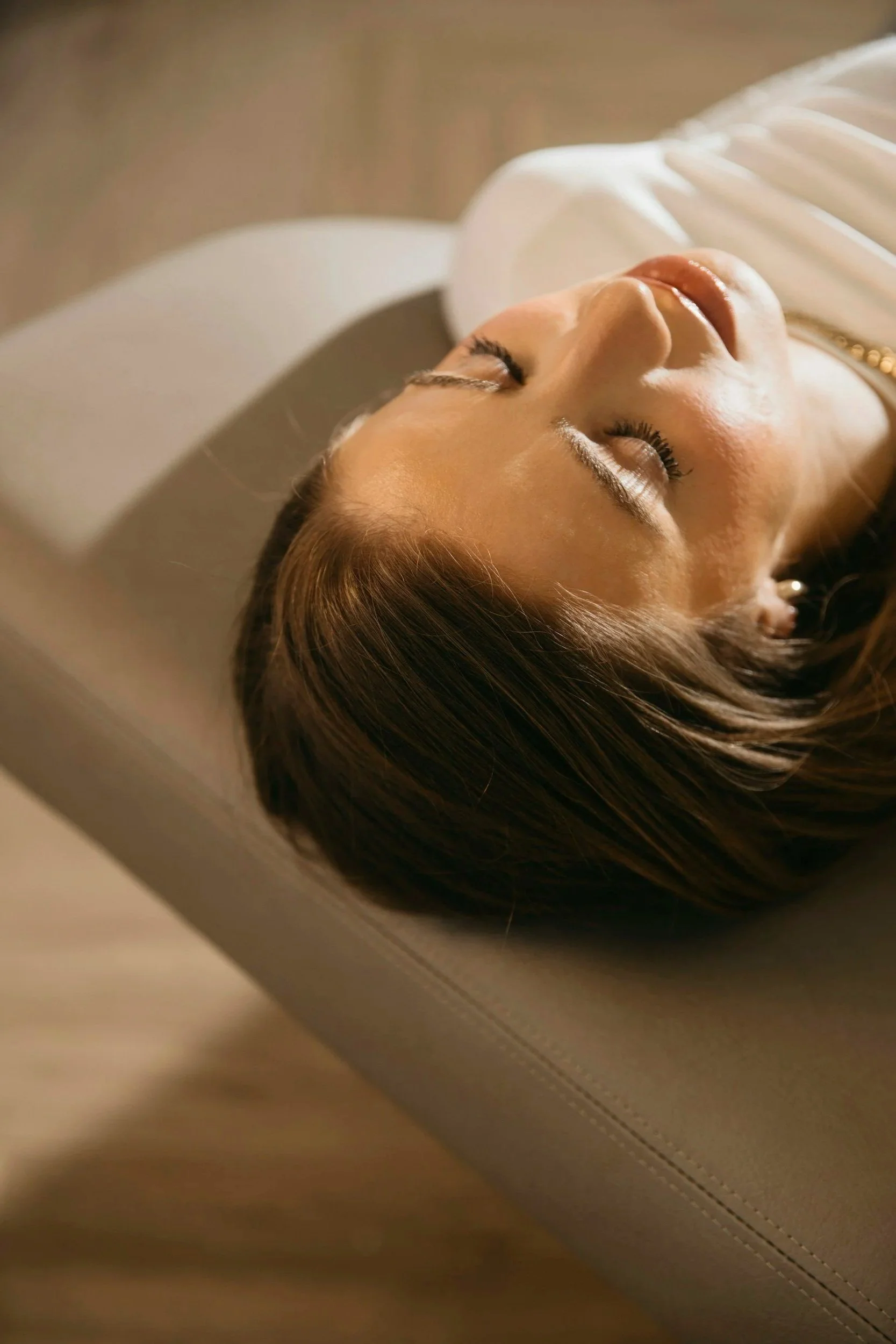A woman with brown hair and closed eyes, lying on a beige couch in a relaxed state.