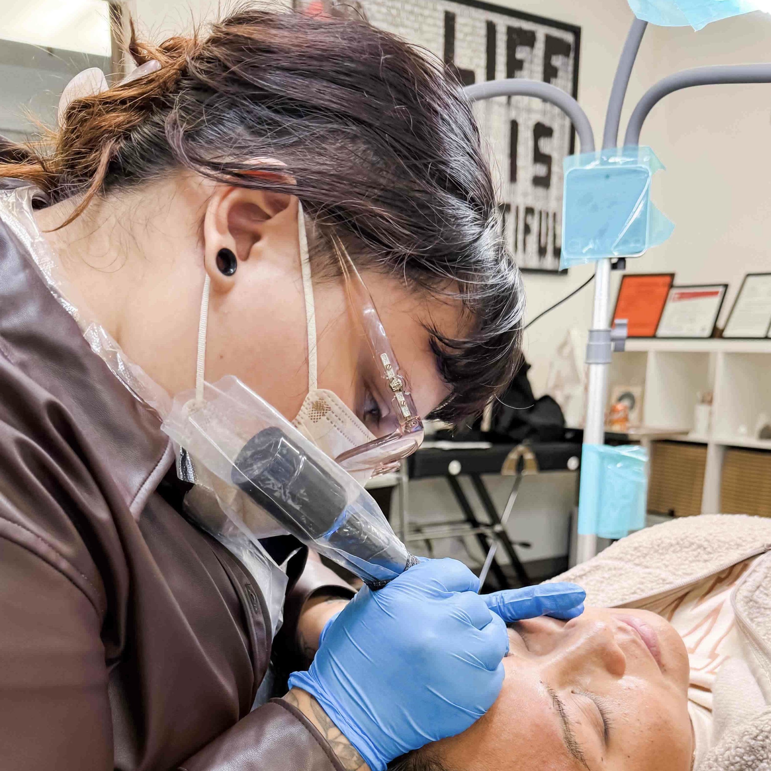 A permanent makeup tattoo artist in PPE protective gear; including gloves, mask, apron and glasses, is creating a realistic nano brow tattoo on a person's eyebrows in a professional studio setting.