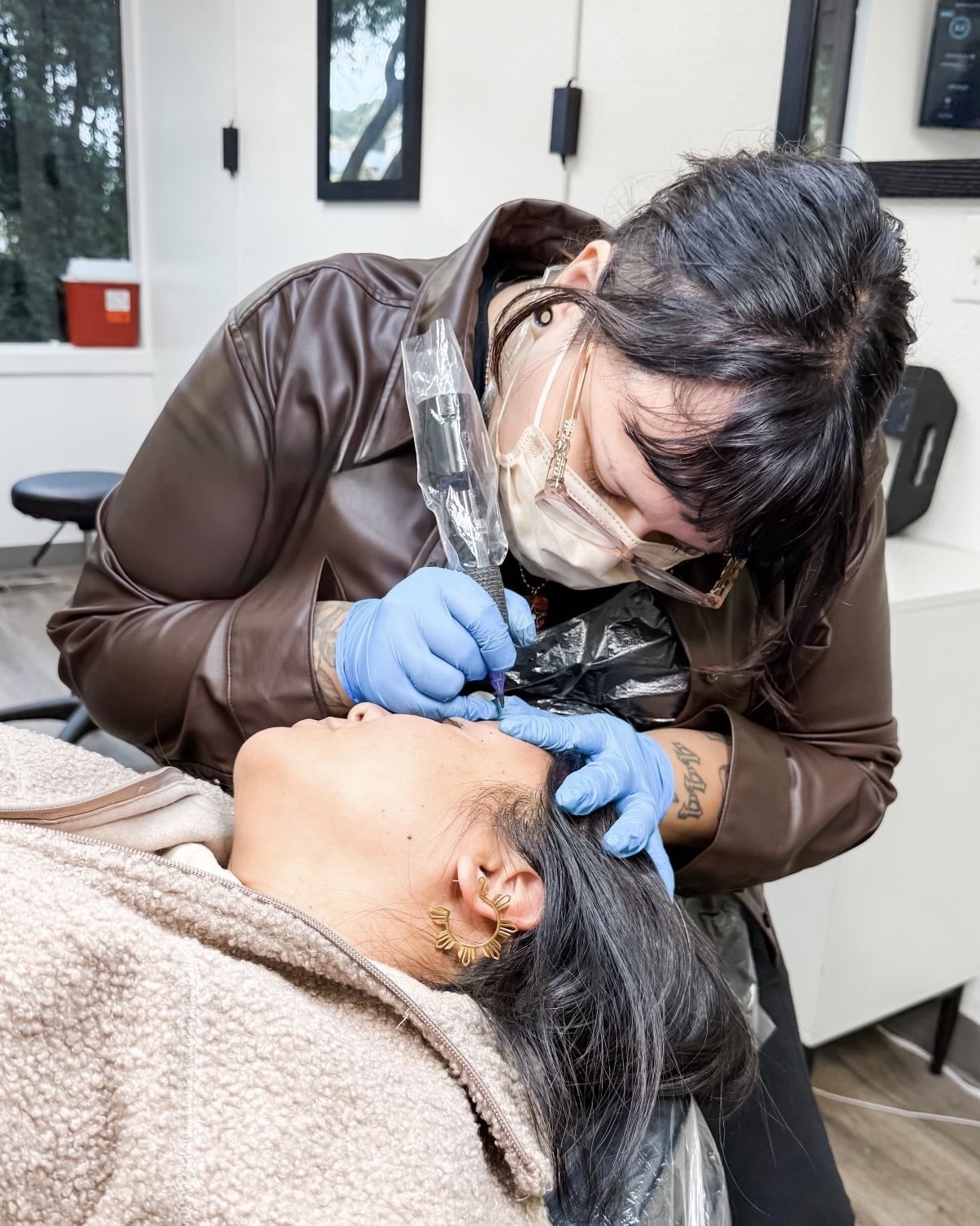 Permanent makeup tattoo artist performing a nano brow machine hair strokes tattoo on a woman's eyebrows in a professional studio, wearing PPE protective equipment like a mask, apron, glasses and gloves.