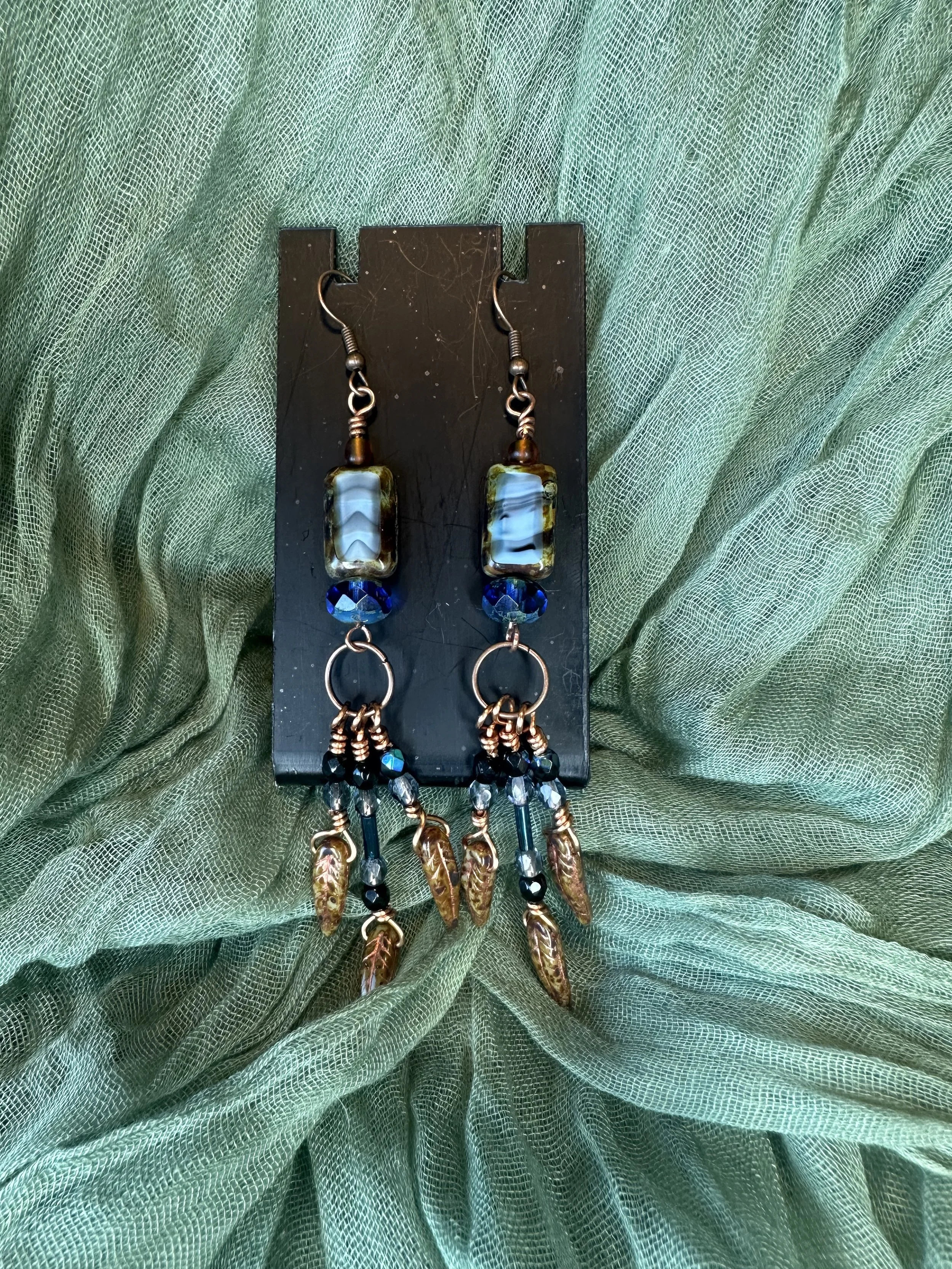 A pair of beaded dangle earrings with blue, black, and gold accents displayed on a black card, set against a flowing green fabric background.