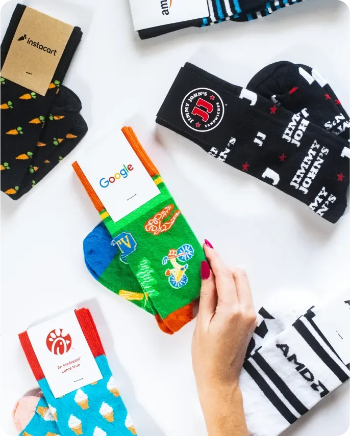 Colorful socks with various designs, including hot dogs, ice cream, and the Google logo, placed on a white surface. Some socks are labeled with brand tags, and a hand with painted nails is holding one of the socks.