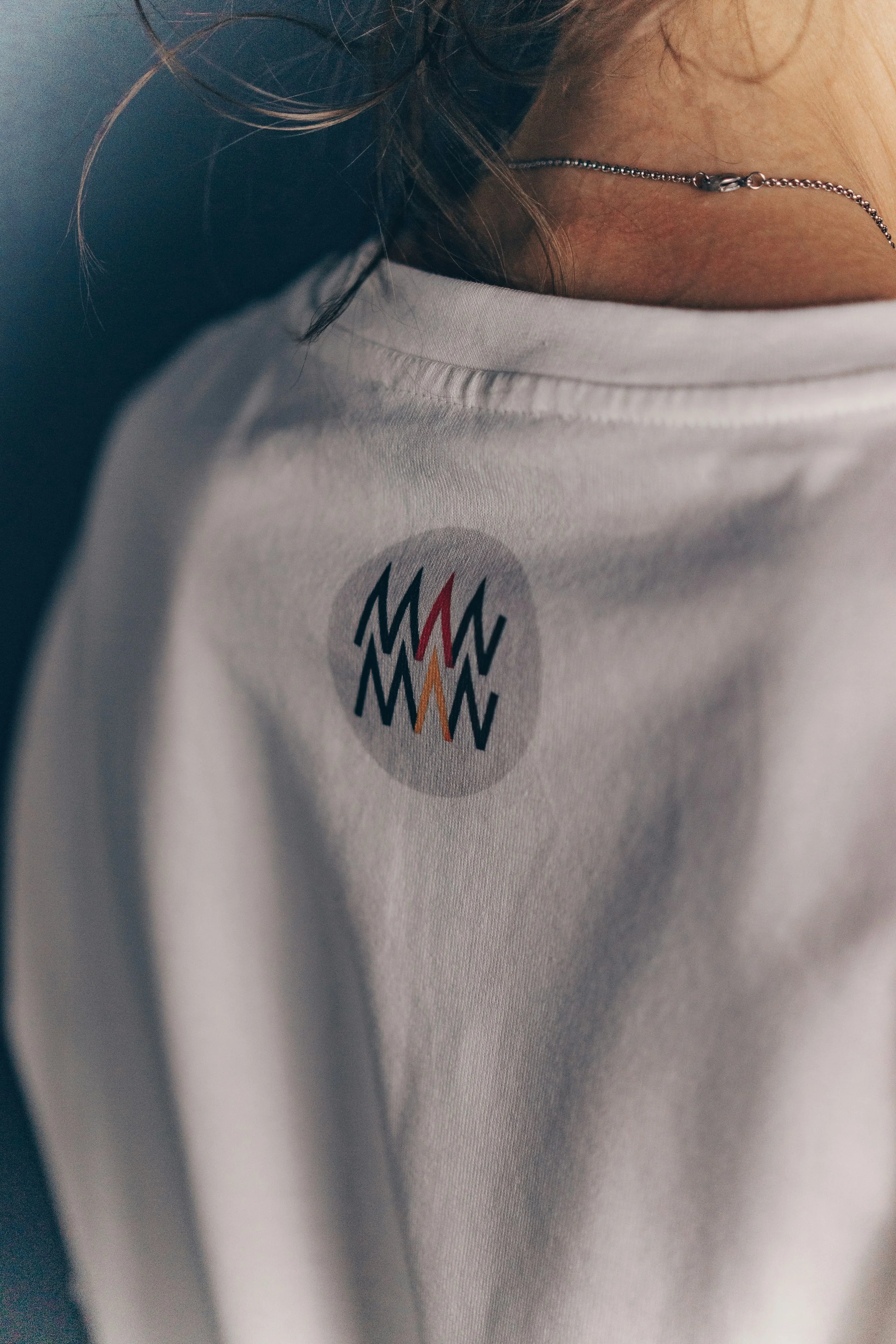 Close-up of a person's shoulder and neck wearing a white T-shirt with a colorful logo, showing a decorative necklace.