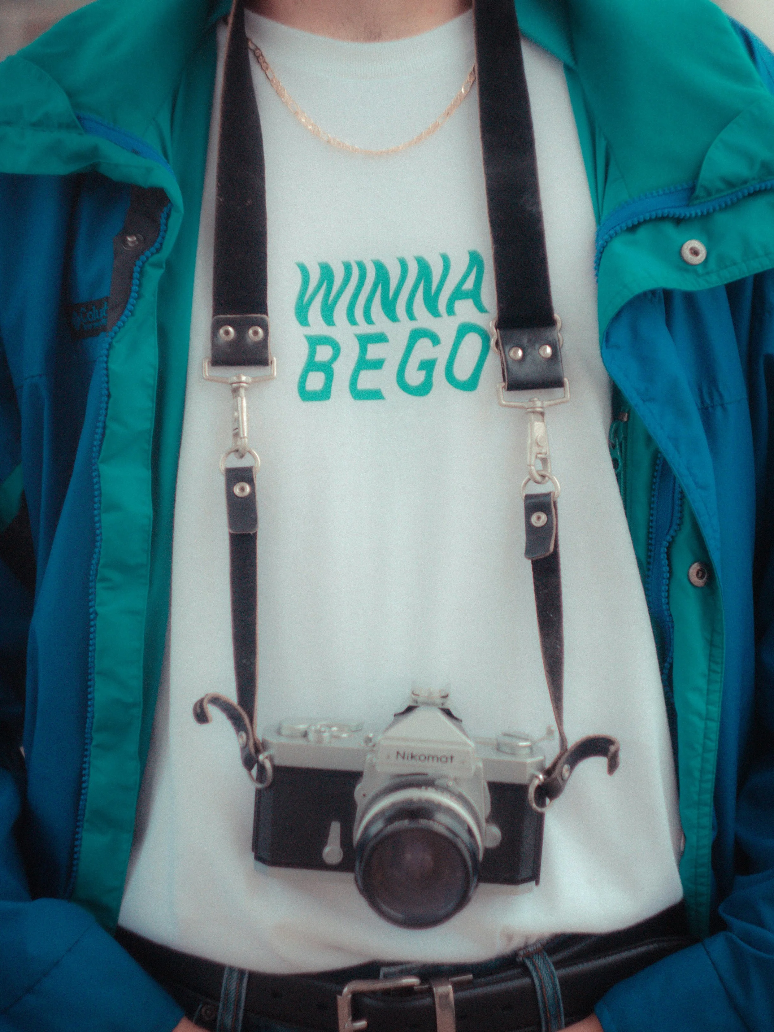 Person wearing a teal and blue jacket with a white T-shirt that says 'WINNA BEGO,' and a Nikon camera hanging from a strap around their neck.