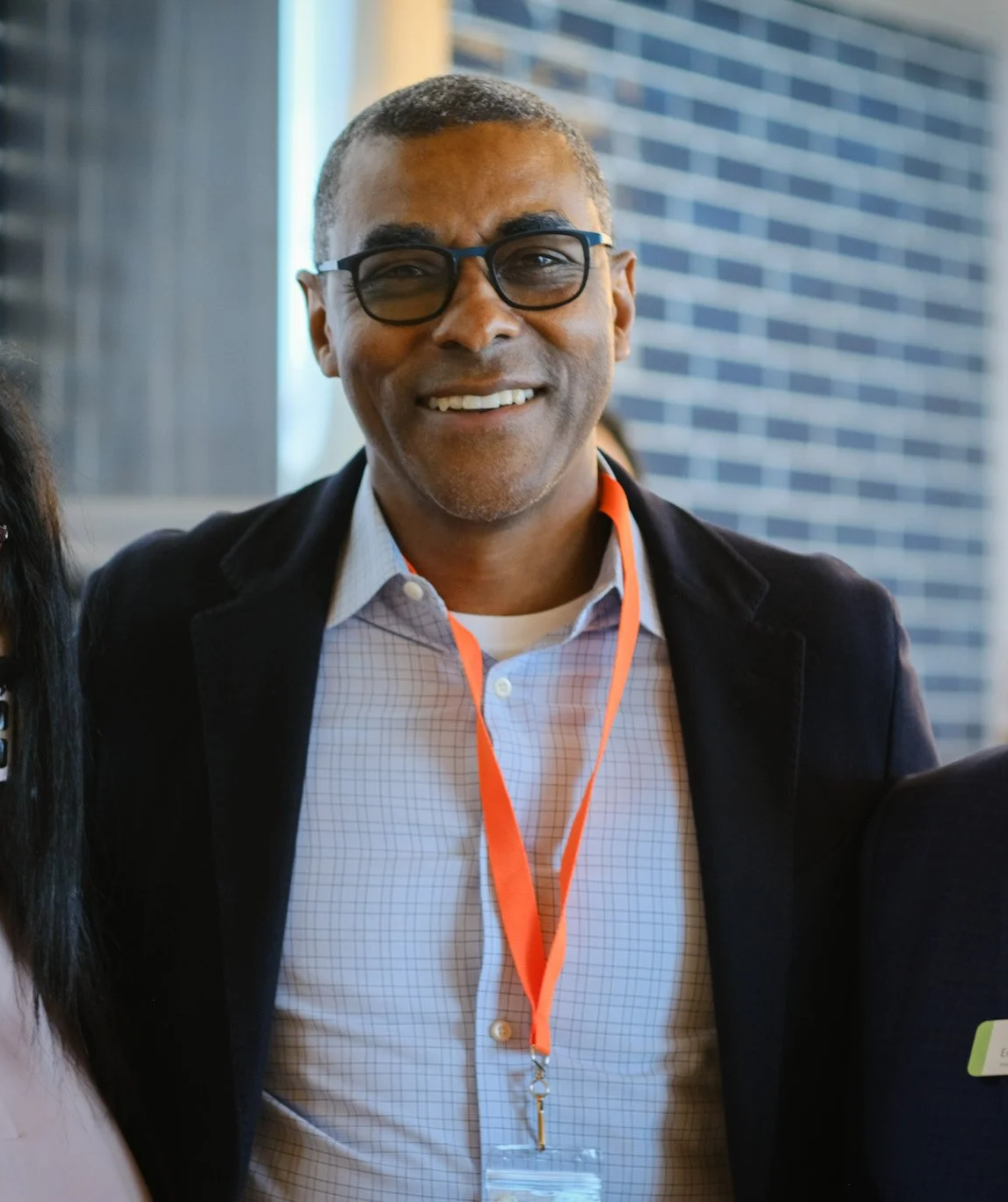 A smiling man with short gray hair and glasses wearing a black jacket and a lanyard with a badge at a professional event, with a brick building in the background.