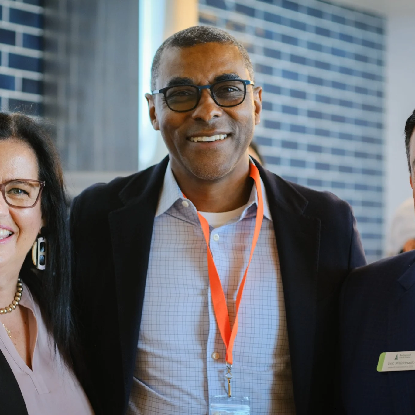 Three people smiling at a professional event, with a man in glasses at the center wearing a safety badge around his neck.