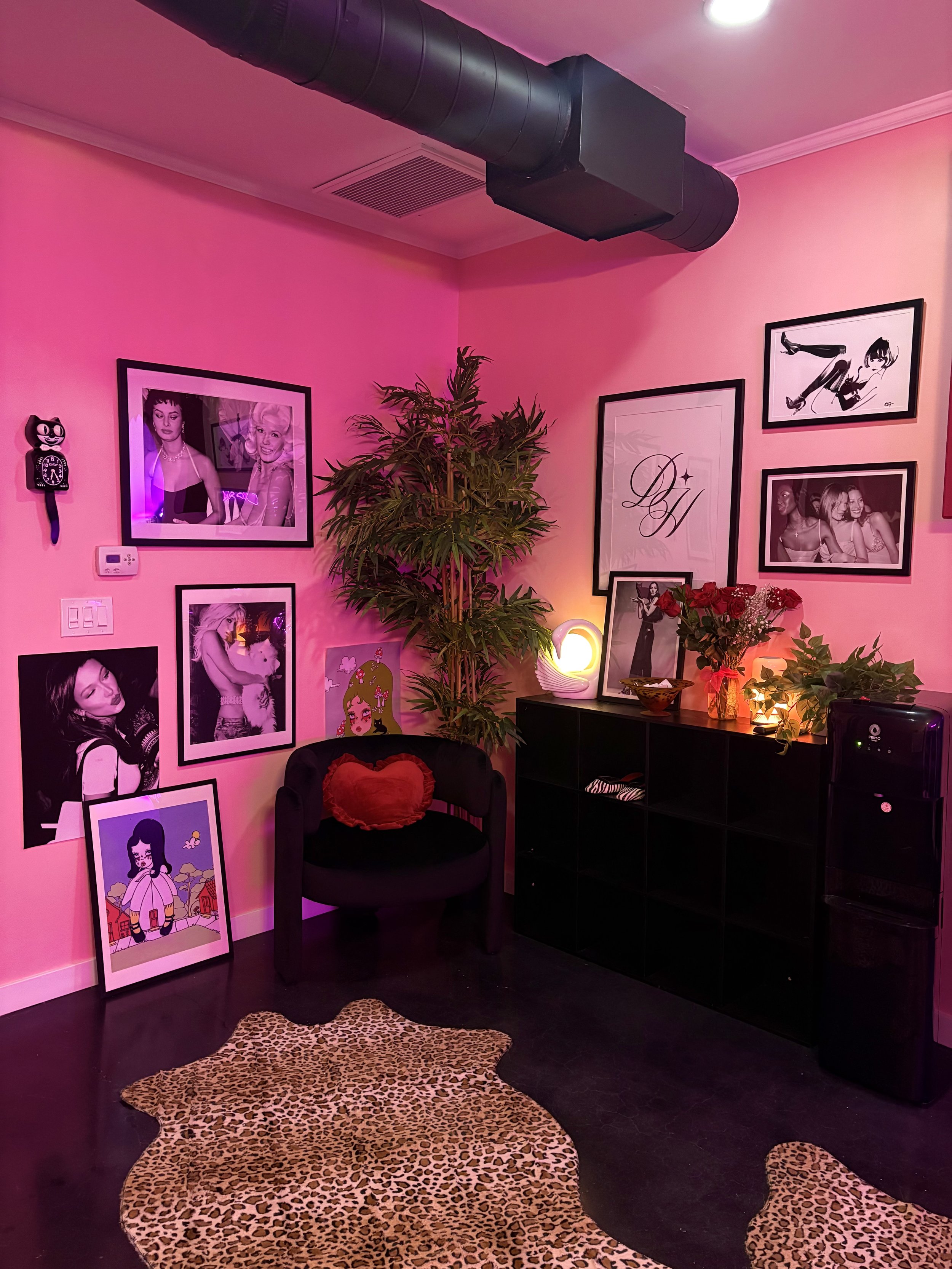 A pink-themed room with framed photographs and artwork on the wall, a potted plant, small lamp, and roses on a black shelf, a chair with a red heart-shaped pillow, a leopard print rug, and a water cooler.