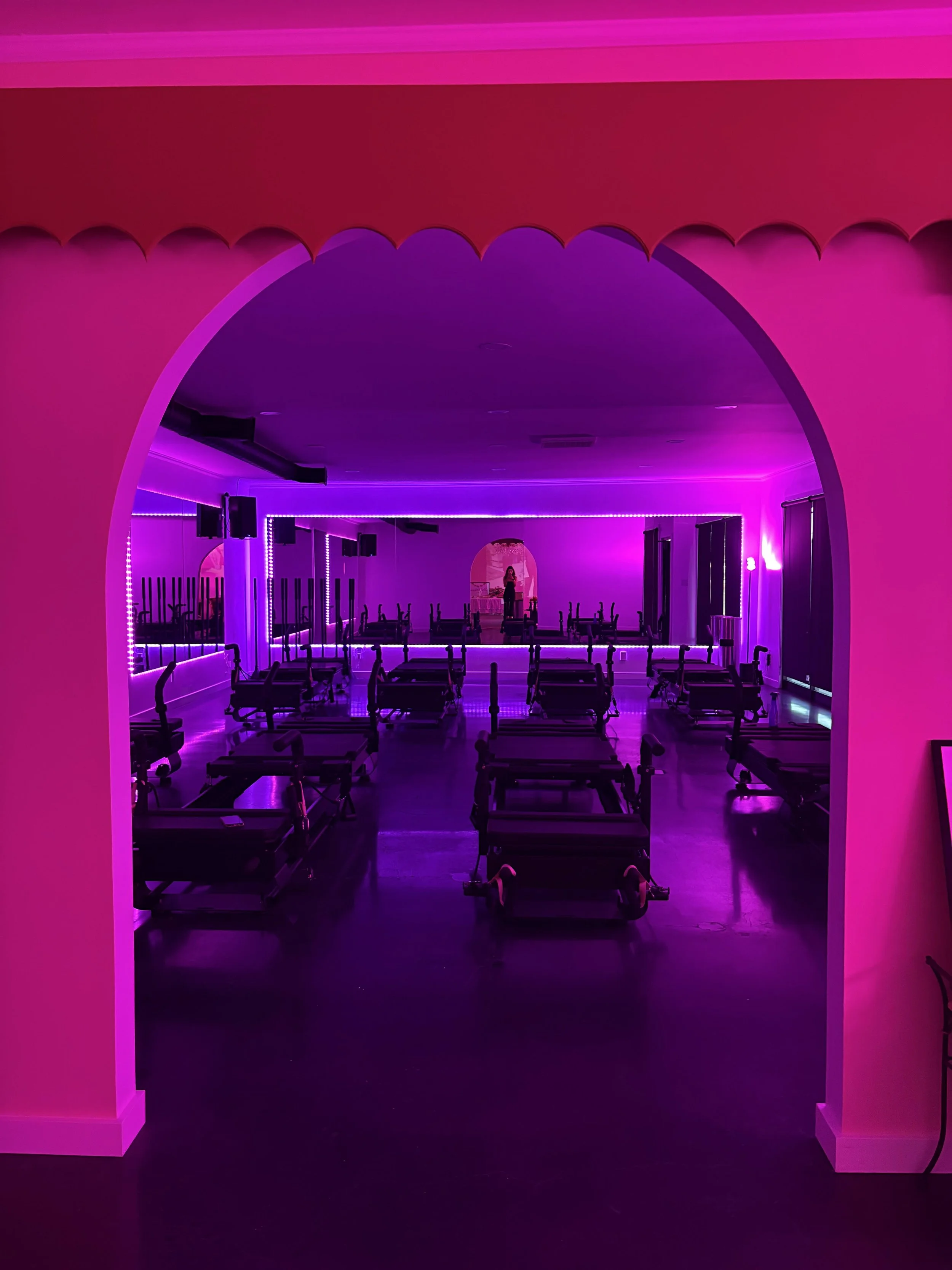 A dimly lit room with purple LED lighting and exercise equipment, including Pilates reformer machines, arranged in rows. A person is standing on a stage at the back of the room.
