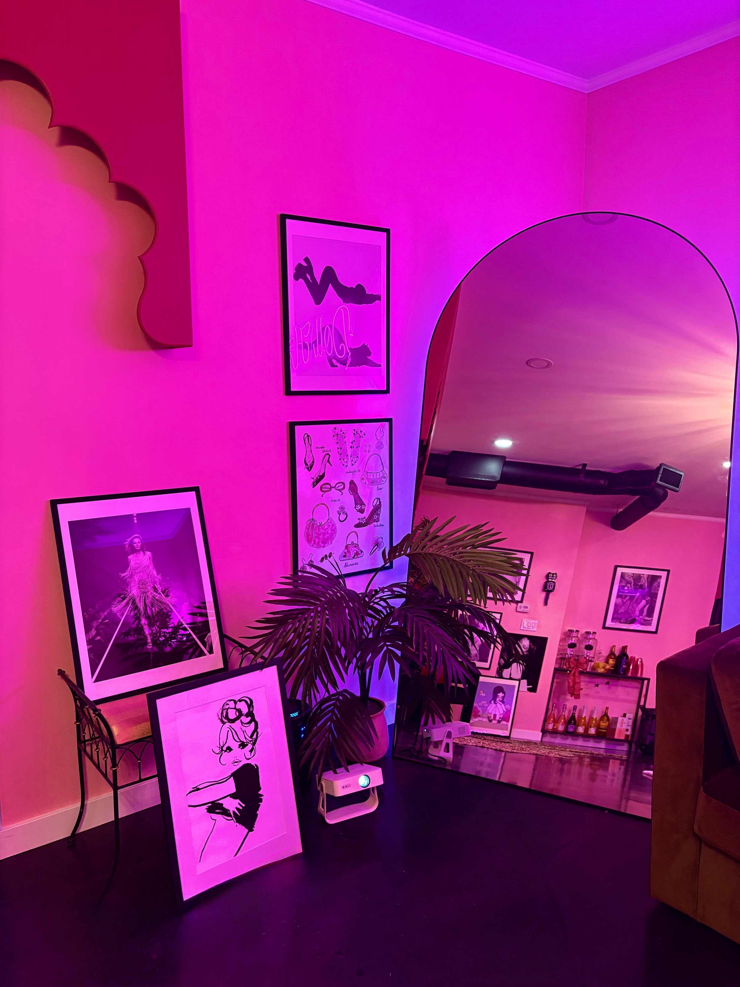 A cozy indoor space decorated with pink and purple lighting. Framed artwork and photographs leaning against the walls and on the floor, including a drawing of a woman and fashion illustrations. A large potted plant adds greenery. The space features an arched wall opening, a mirror, and a small bar area with bottles and glassware.
