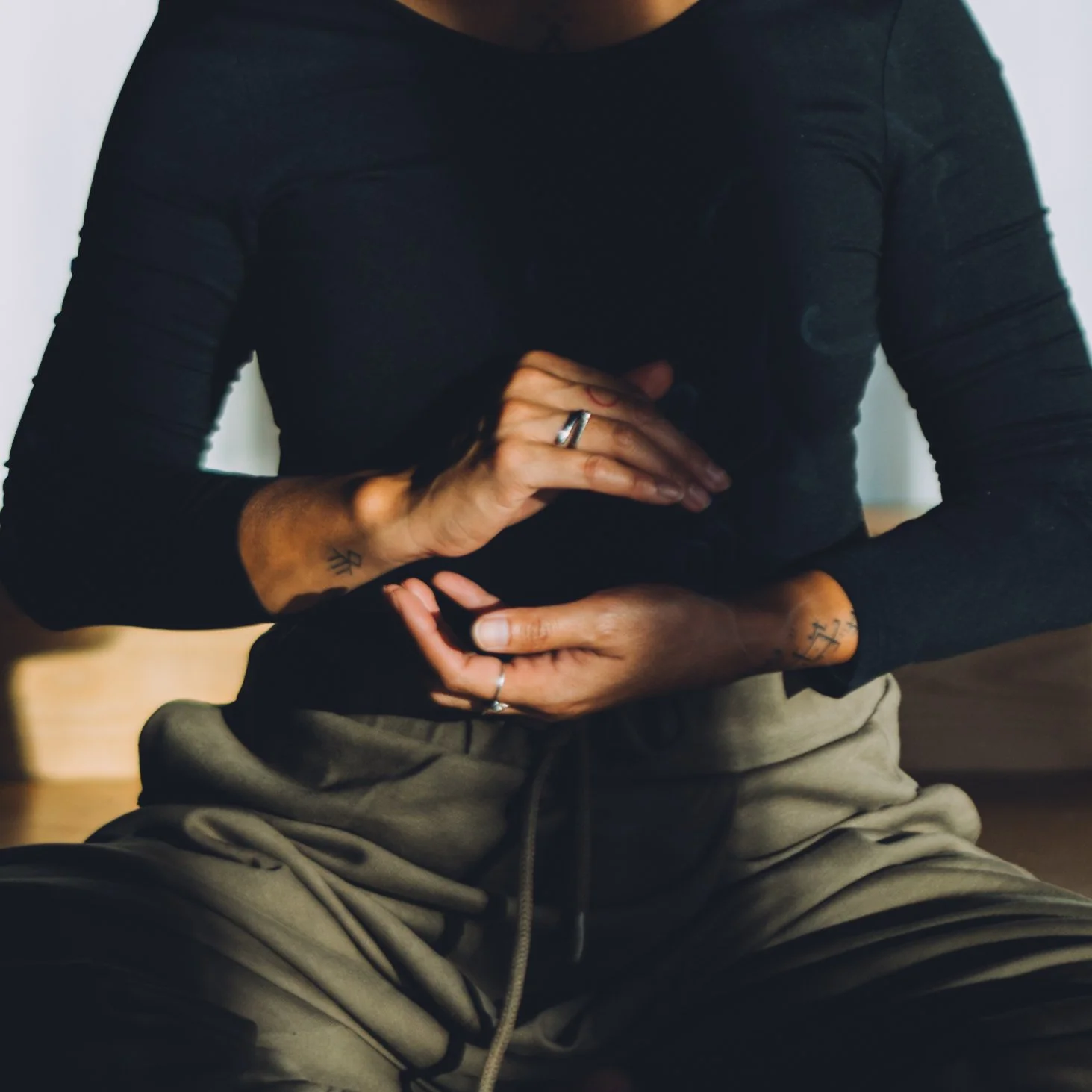 Une personne assise, portant un haut noir en tissu extensible et un pantalon beige, avec des tatouages visibles sur les mains et les bras.