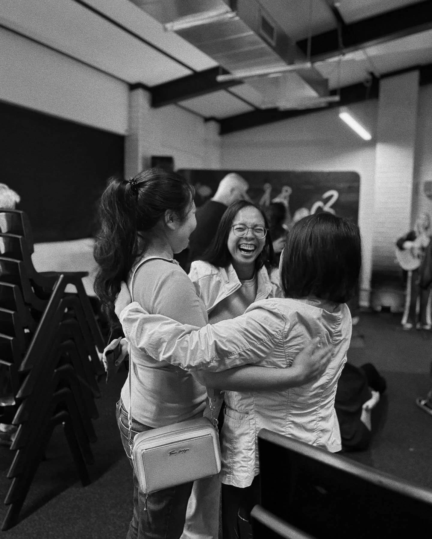 Three women are joyfully embracing each other in a room with other people in the background. One woman is wearing glasses and laughing, while the other two have their backs to the camera.