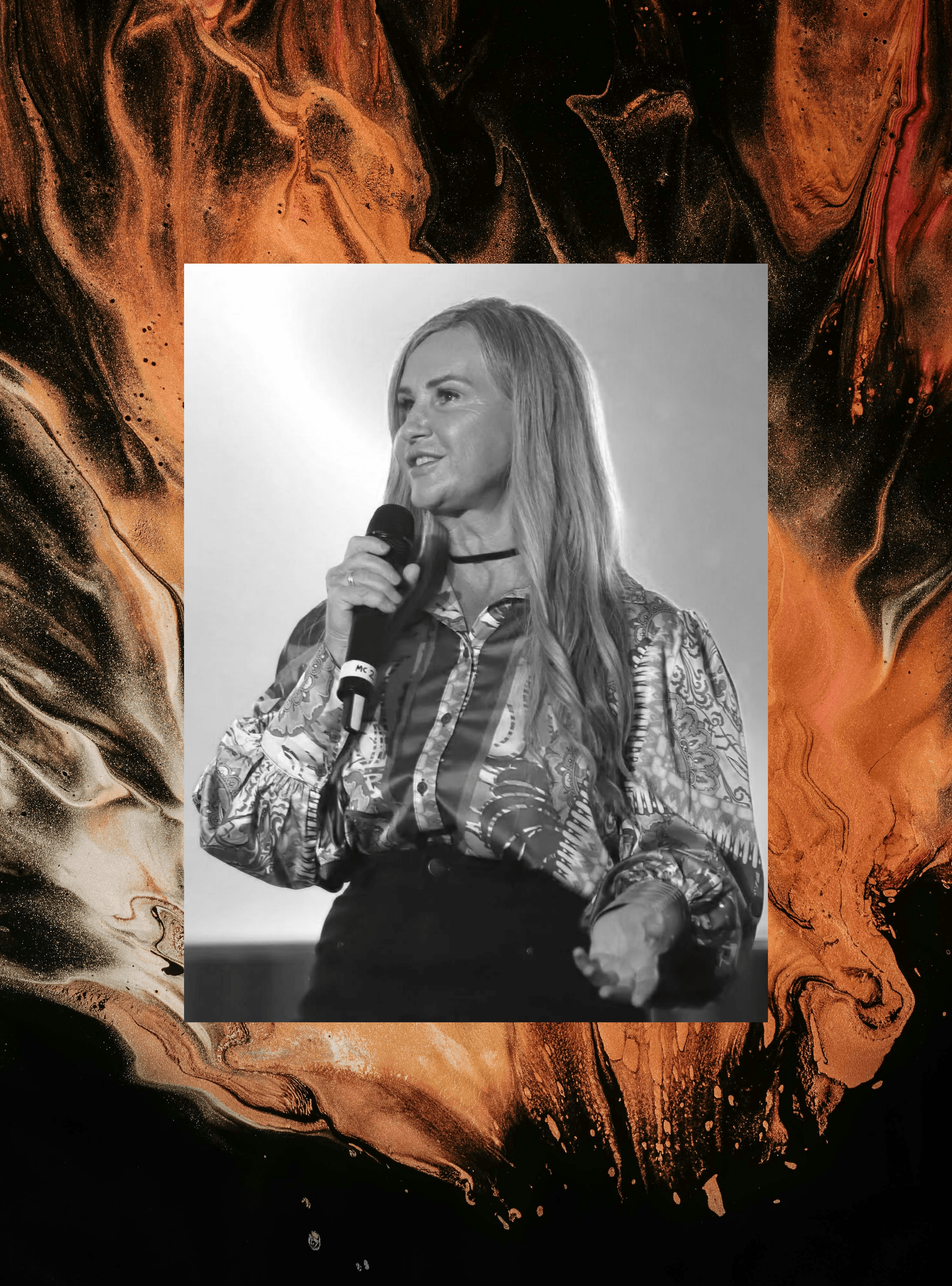 Black and white photo of a woman holding a microphone, speaking or singing, with long hair, wearing a patterned blouse.