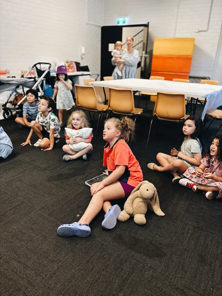 Children sitting on the floor in a room, watching something, with a stuffed bunny toy near one girl, and an adult woman holding a young child in the background.