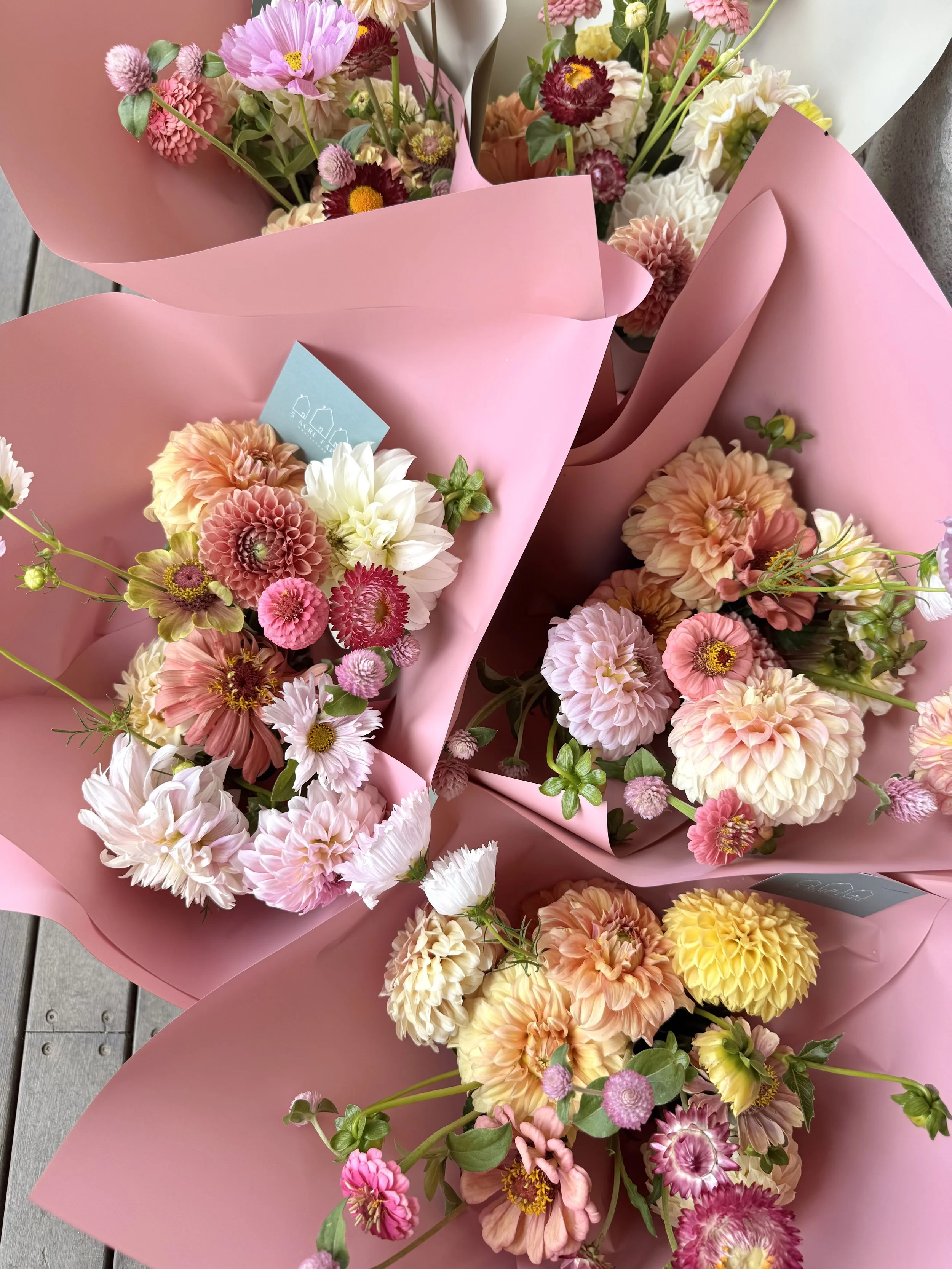 Several colorful flower bouquets wrapped in pink paper, including dahlias, daisies, and other mixed flowers, arranged on a wooden surface.