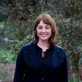 A smiling woman with shoulder-length brown hair and bangs, wearing a dark blazer, standing outdoors with blurred greenery in the background.