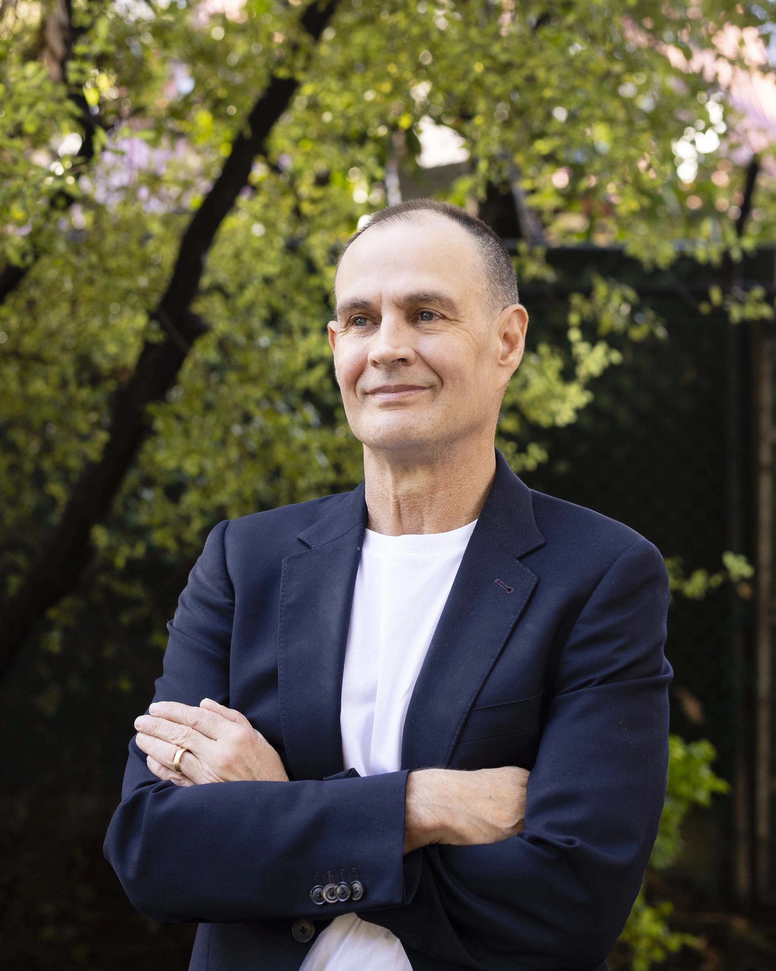 A man with short, dark hair dressed in a navy blazer and white T-shirt standing outdoors in front of green trees with cross arms and a slight smile.
