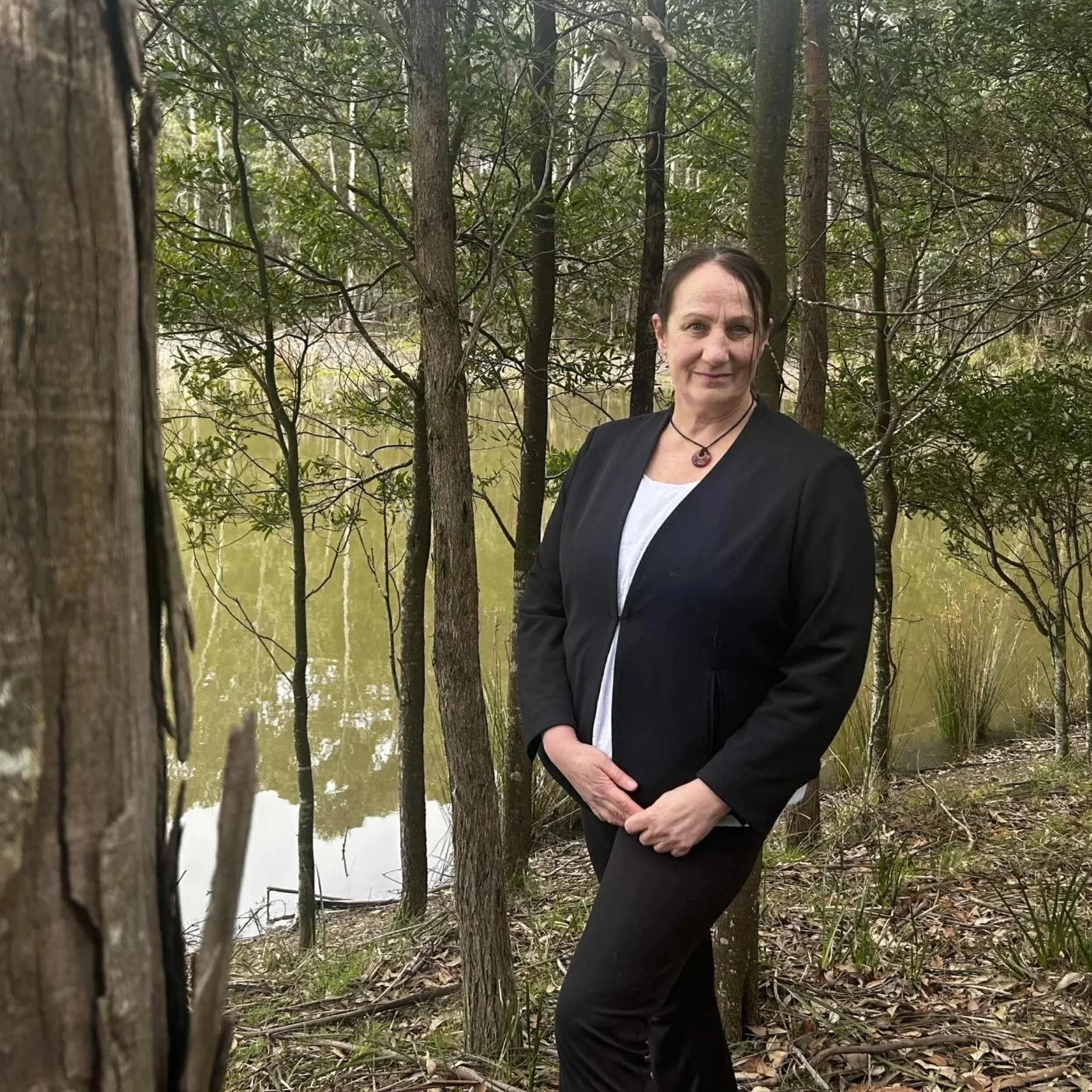 A woman wearing a black blazer and white shirt stands among trees near a pond within a forested area.