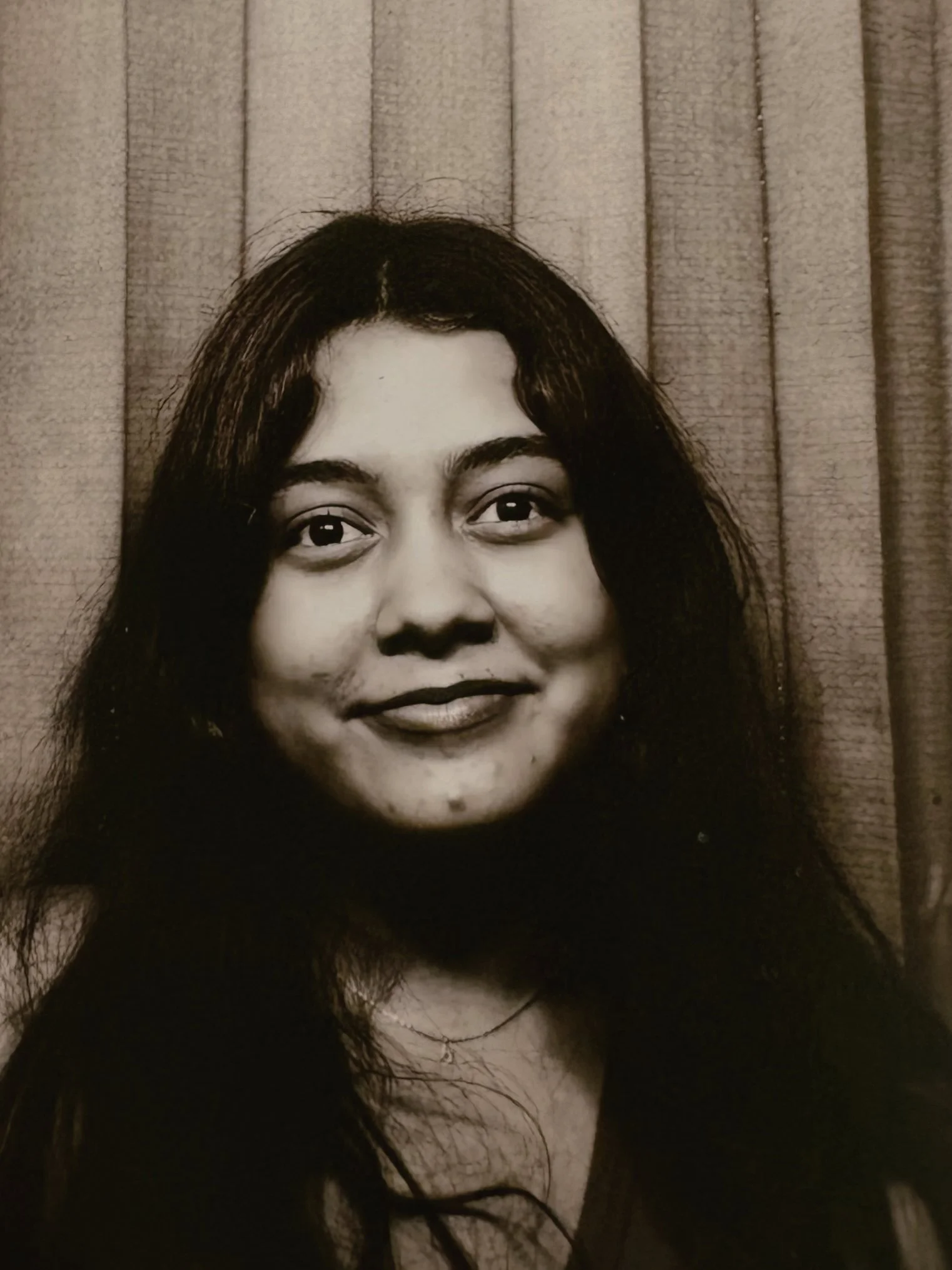 A black and white portrait of a woman with long, dark hair smiling at the camera, standing in front of patterned curtains.