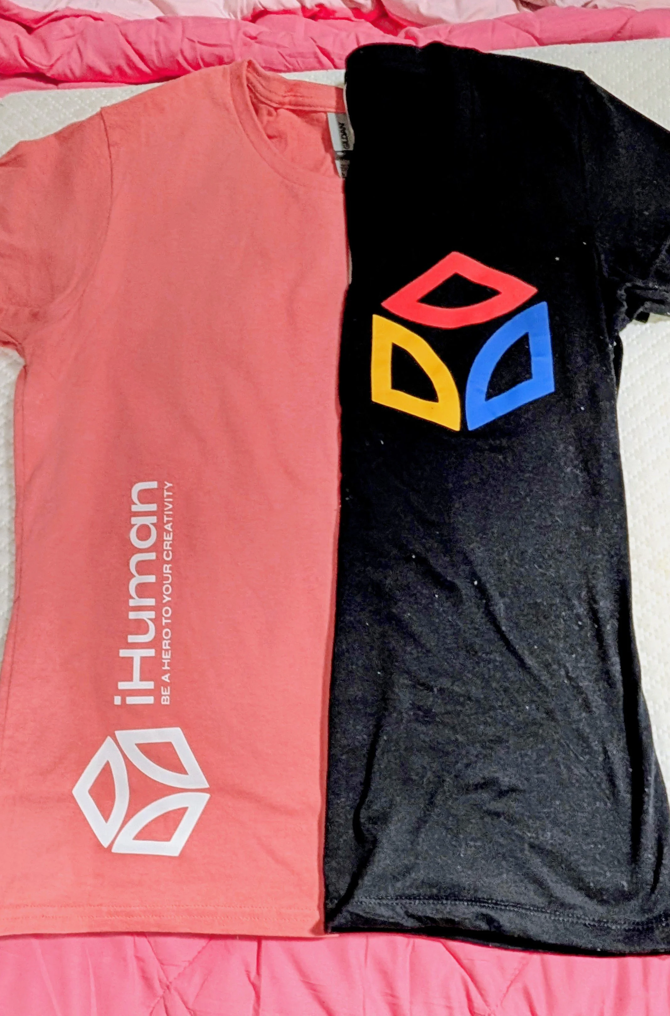 Two T-shirts laid side by side on a pink bedspread; the left one is pink with white and gray text and logo, the right one is black with a colorful geometric logo.